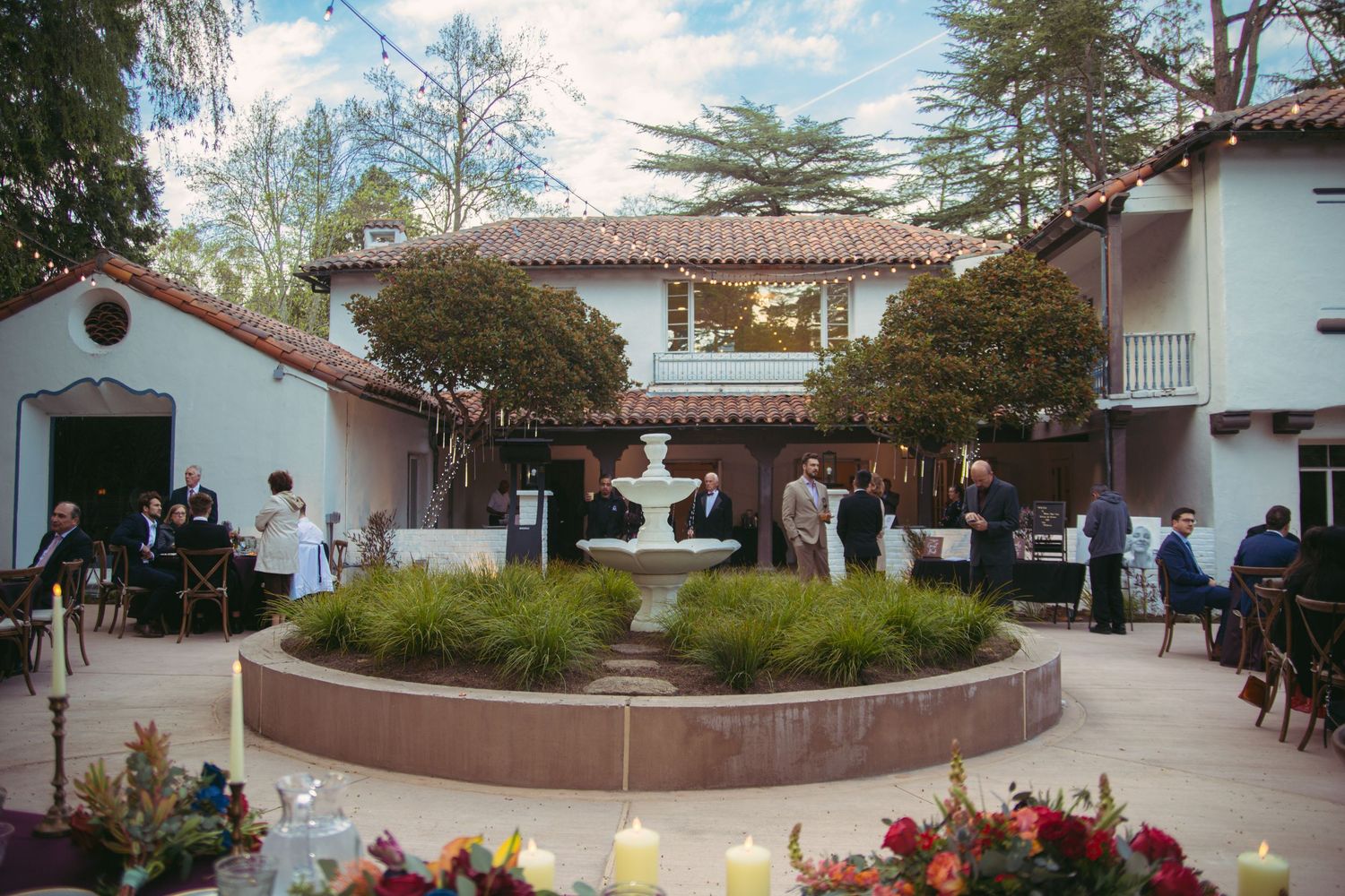 Outdoor wedding venue courtyard with fountain, white Spanish-style building, and guests mingling.