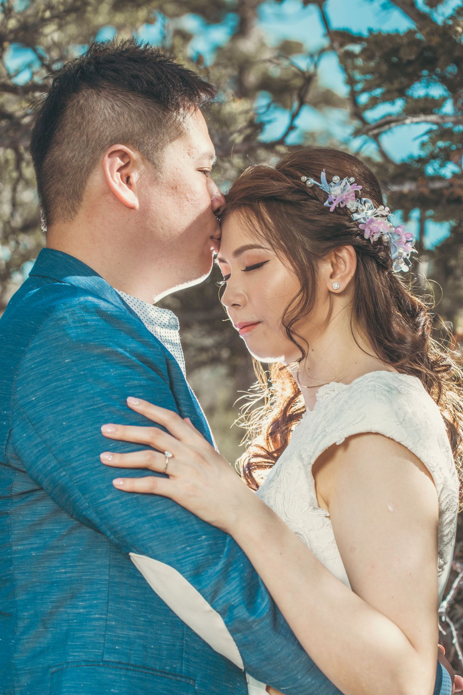A groom in a blue suit kisses his bride wearing a floral crown outdoors among trees.