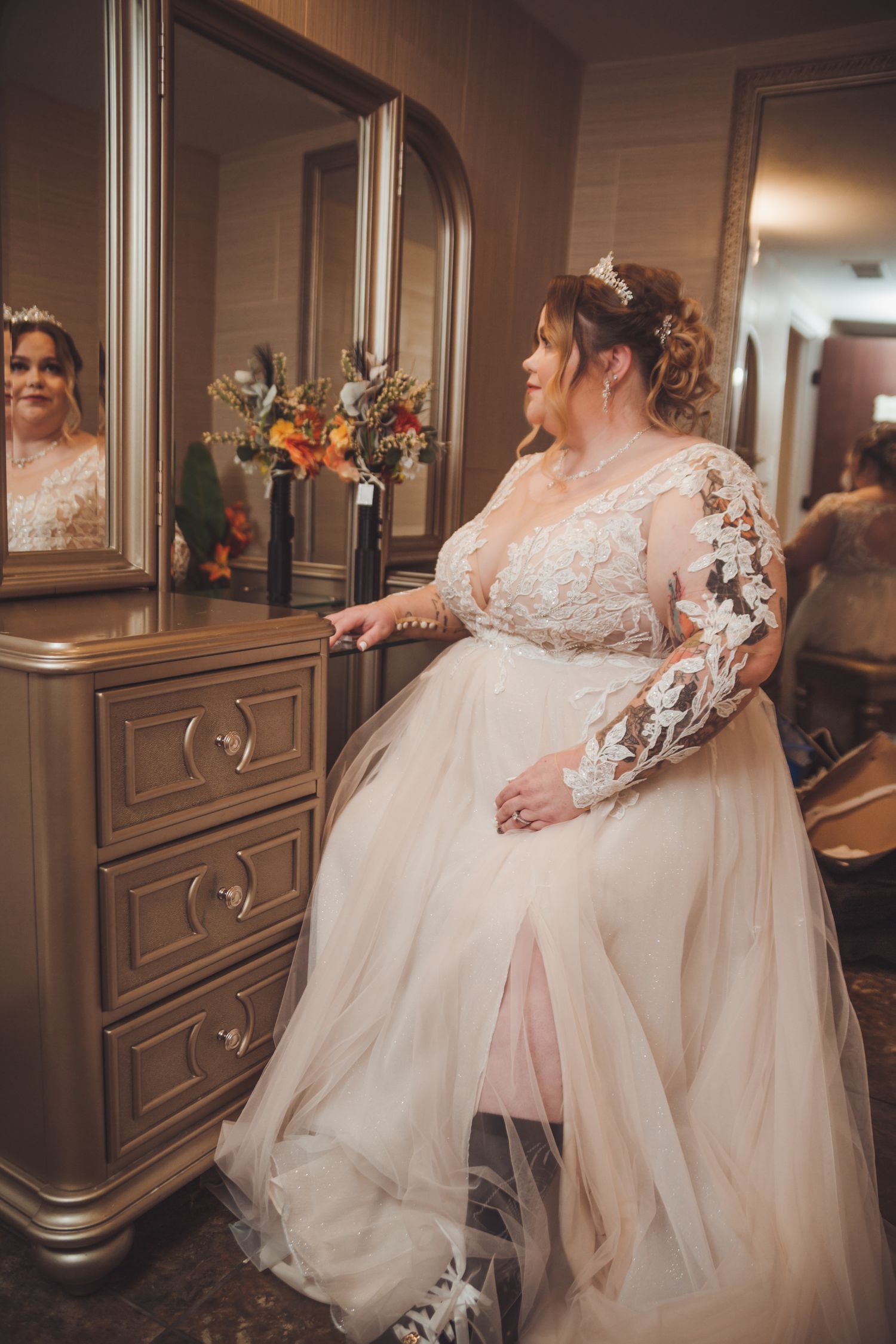 Bride in lace long-sleeve wedding gown with tulle skirt stands by dresser mirror in bridal suite.
