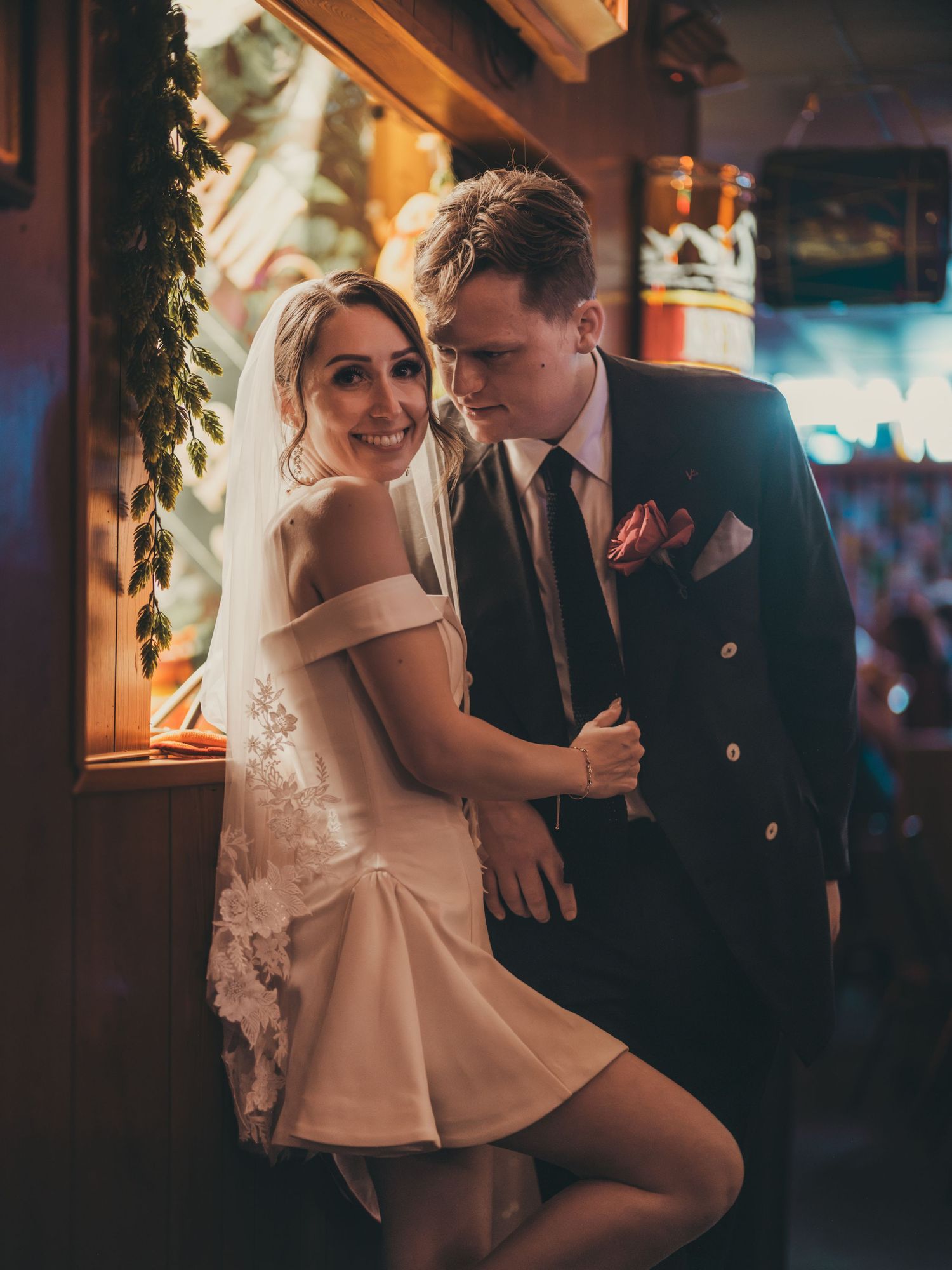 Bride in short white lace dress and veil poses with groom in dark suit at a warmly lit wedding venue bar.
