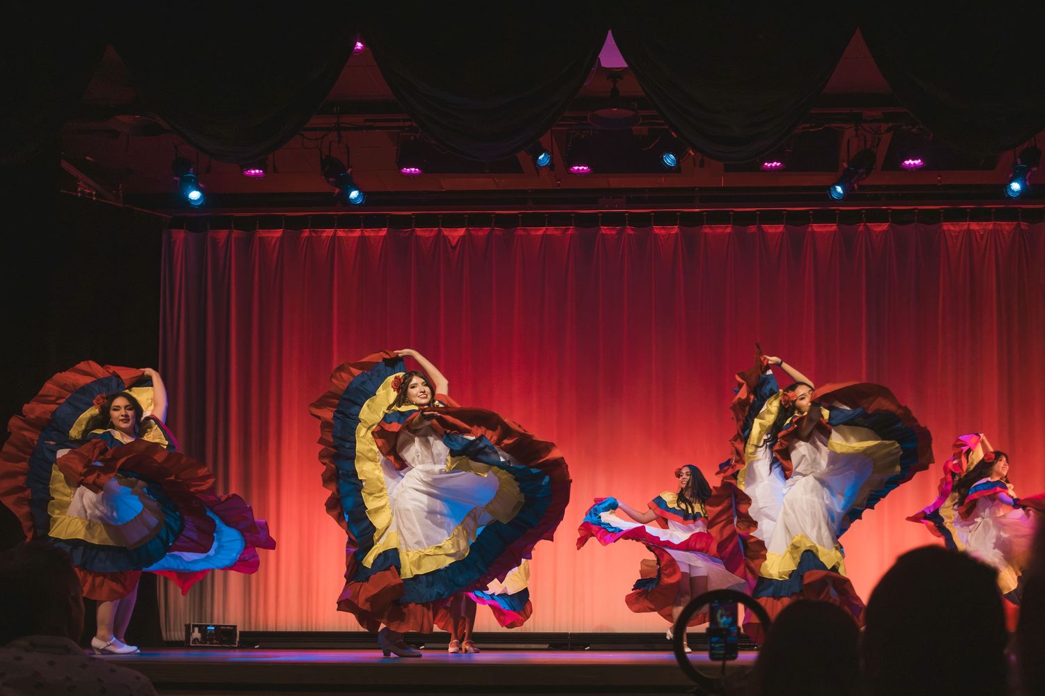 Colorful folkloric dancers in flowing skirts perform on a red-curtained stage under vibrant stage lights.