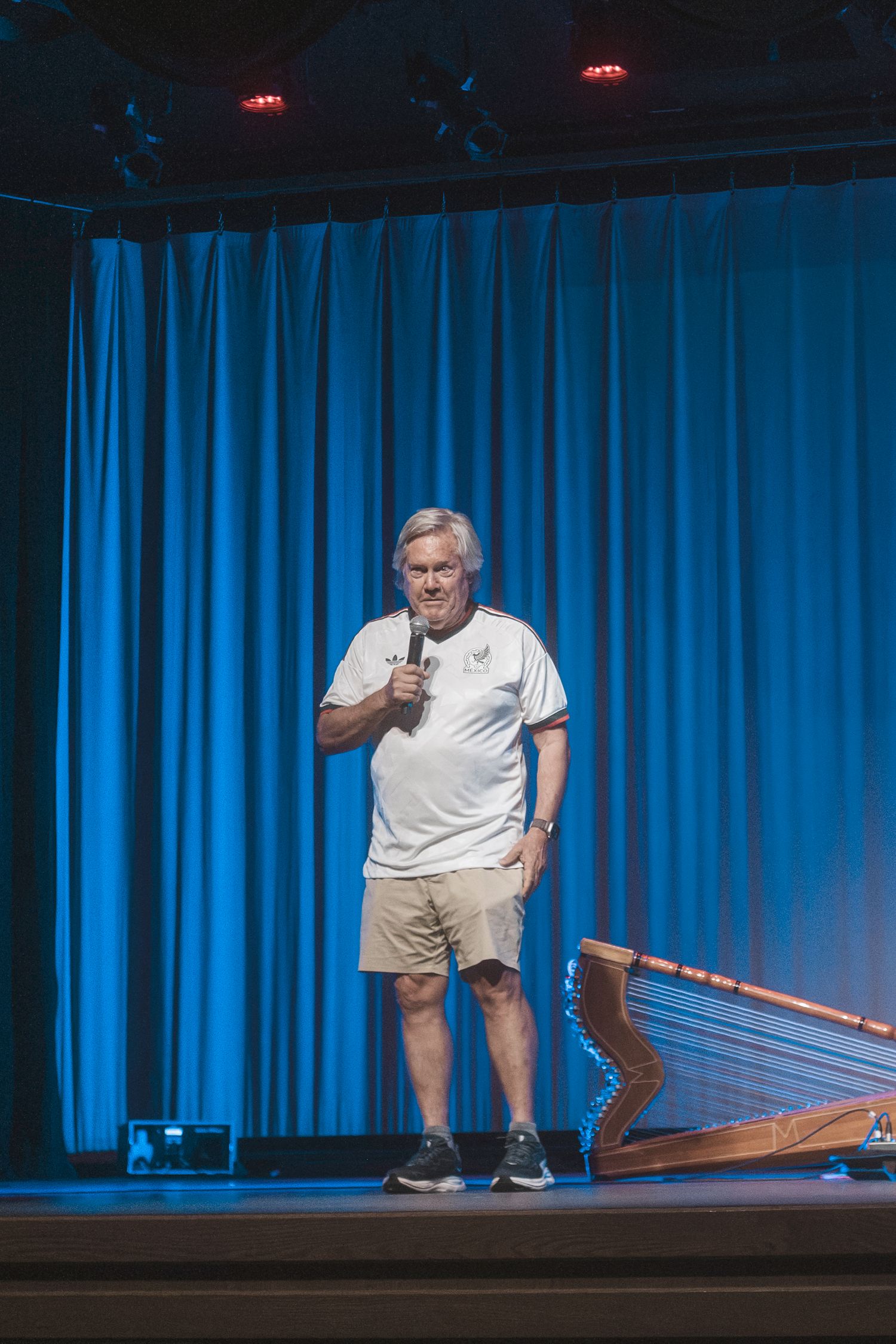 Older man in white t-shirt and khaki shorts holds microphone on stage with blue curtain backdrop.