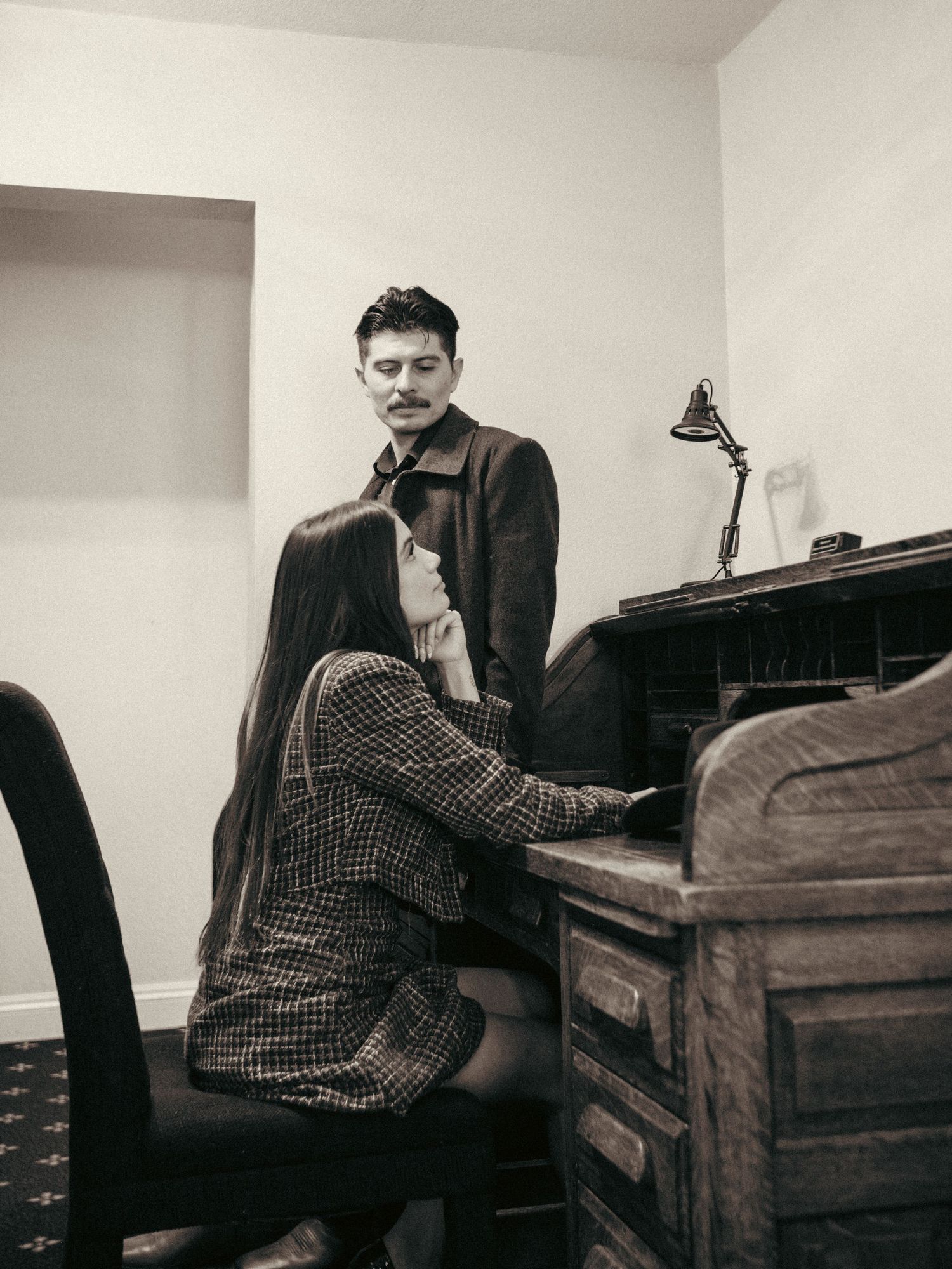 A man stands watching a woman play an antique roll-top desk piano in a vintage black and white photo.