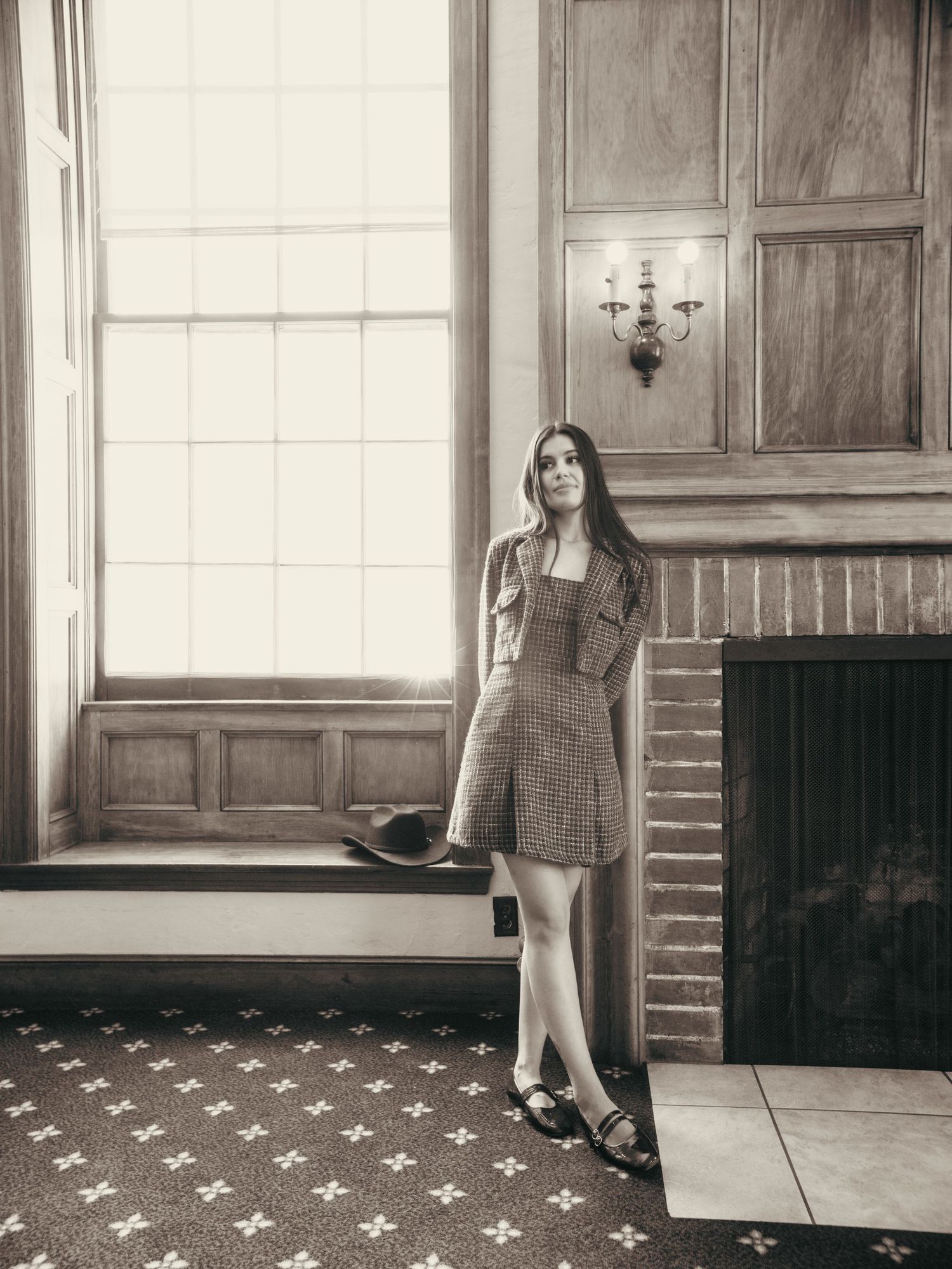 Young woman in plaid mini dress stands by a large window near a fireplace in a vintage wood-paneled room.
