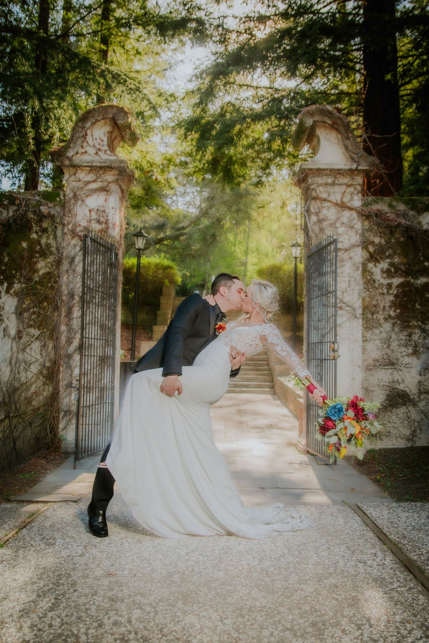 Bride and groom share a romantic dip kiss at an ornate stone garden gate, bride holding colorful bouquet.