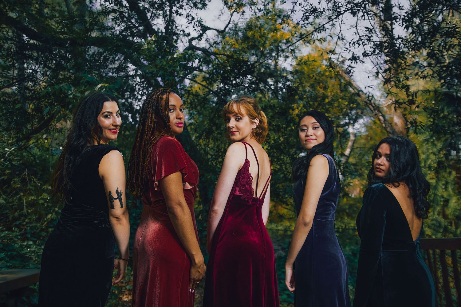 Five women in elegant velvet gowns pose outdoors among lush green trees in a stylish group portrait.