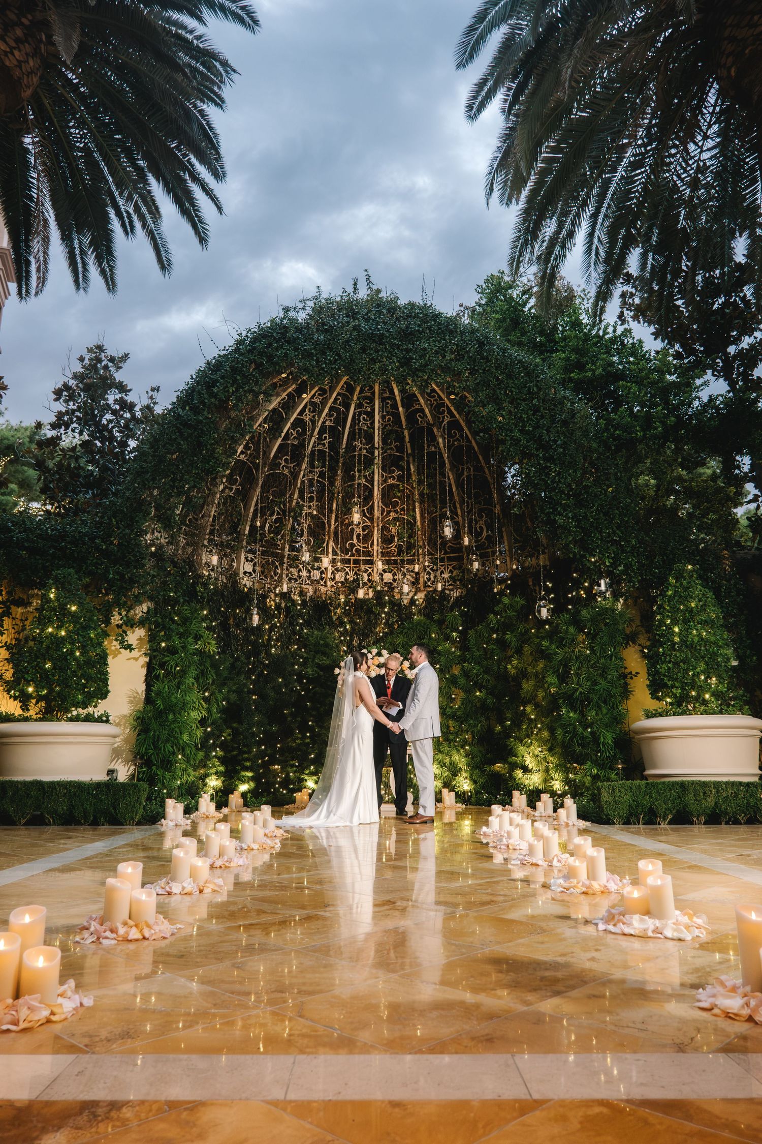 Romantic outdoor wedding ceremony under ivy-covered dome arbor with candles lining the aisle and palm trees at dusk.