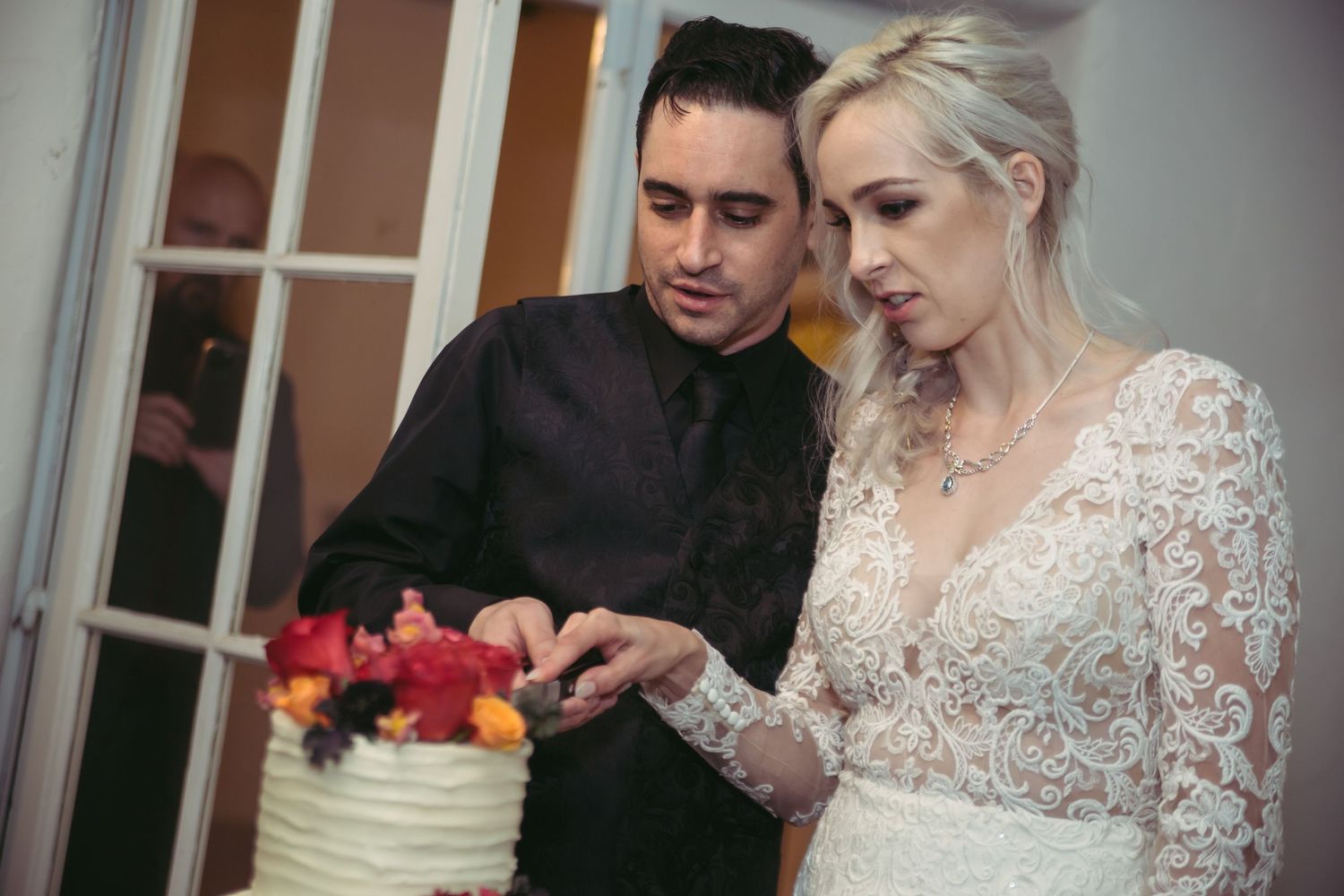 A bride in a lace wedding dress and groom in black suit feed each other wedding cake at their reception.