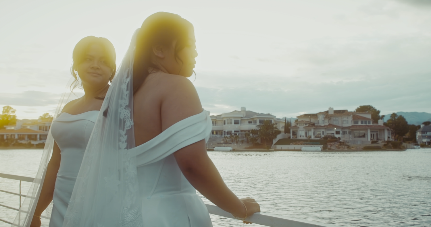 Bride in white gown and veil leans on railing with waterfront homes glowing in golden sunset light.