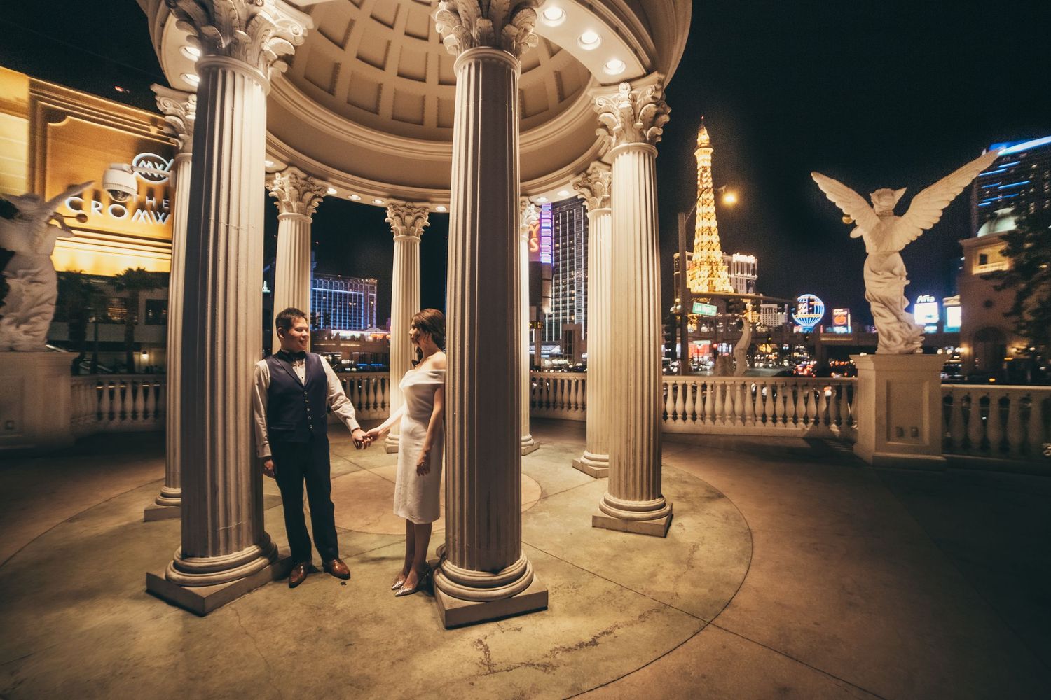 Couple holding hands under Roman columns at night on the Las Vegas Strip with Eiffel Tower replica in background.