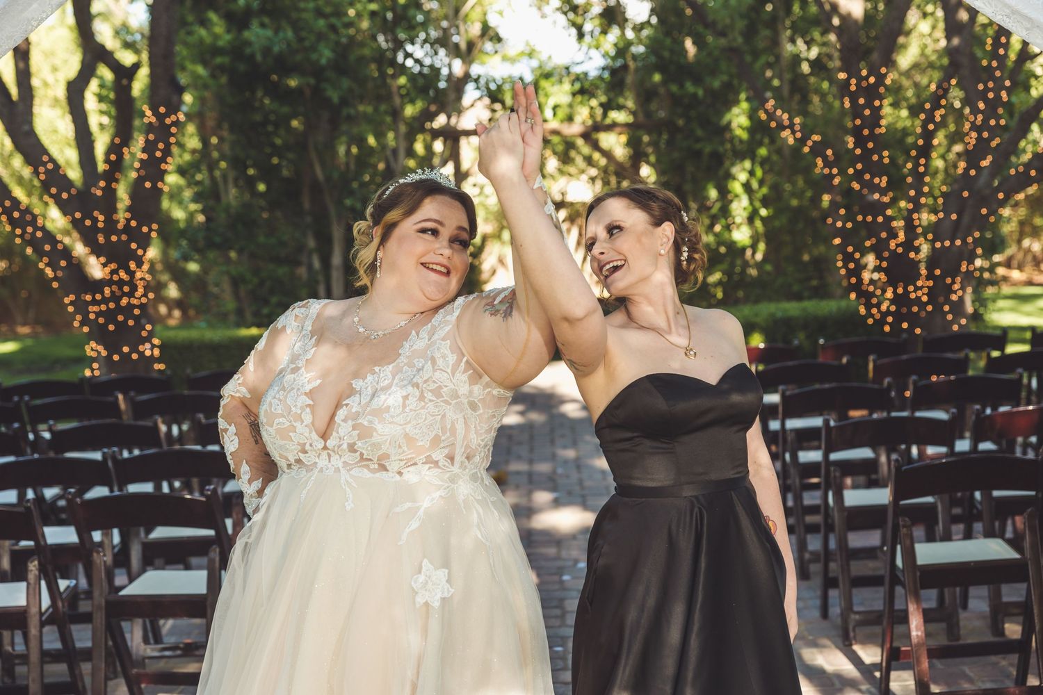 Bride in white lace gown high-fives bridesmaid in black dress at outdoor wedding ceremony venue.