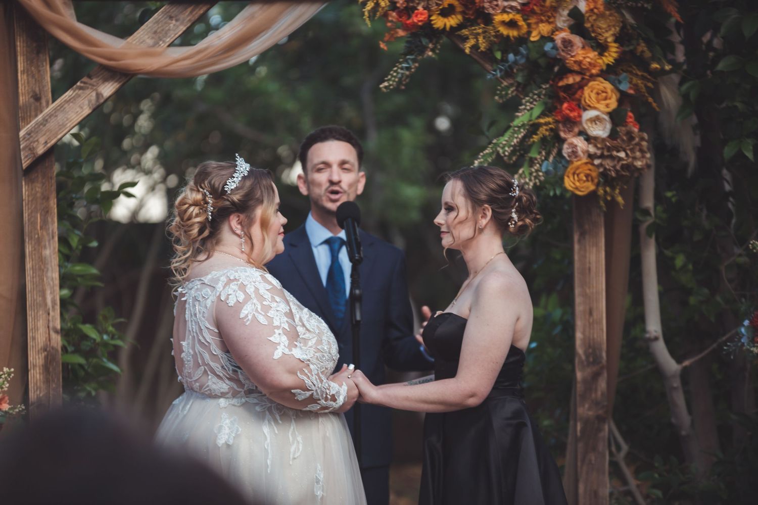 Two brides hold hands at outdoor fall wedding ceremony under rustic wooden arch with sunflowers and colorful blooms.