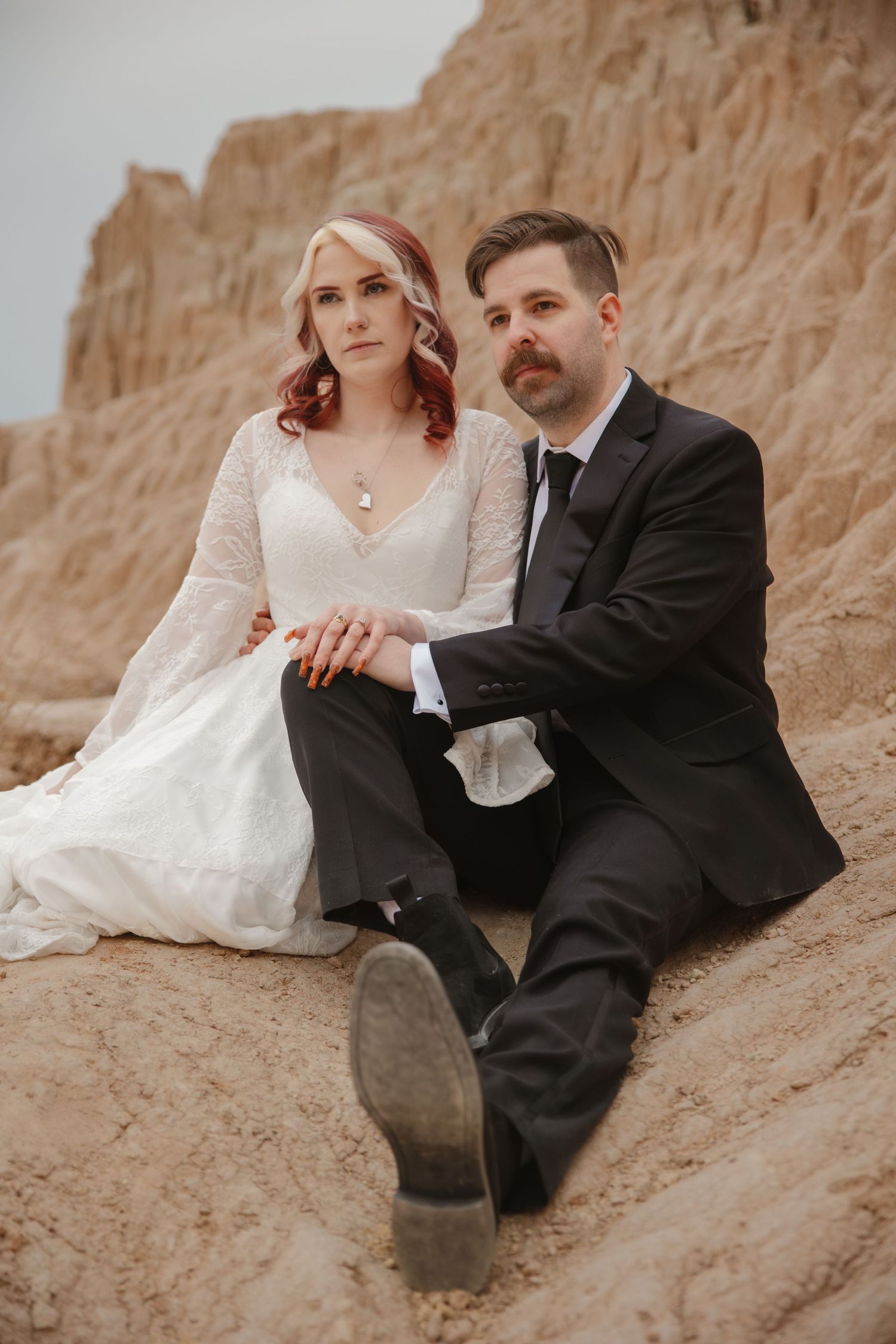 Elegant couple in wedding attire sit on rocky desert landscape for dramatic bridal photo session.
