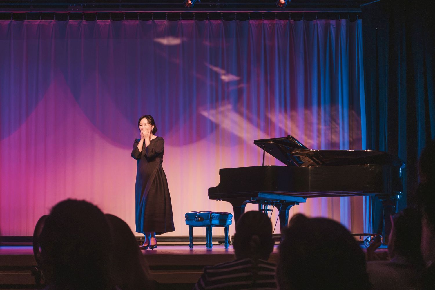 A woman in a black dress performs on stage near a grand piano under pink and blue stage lights.