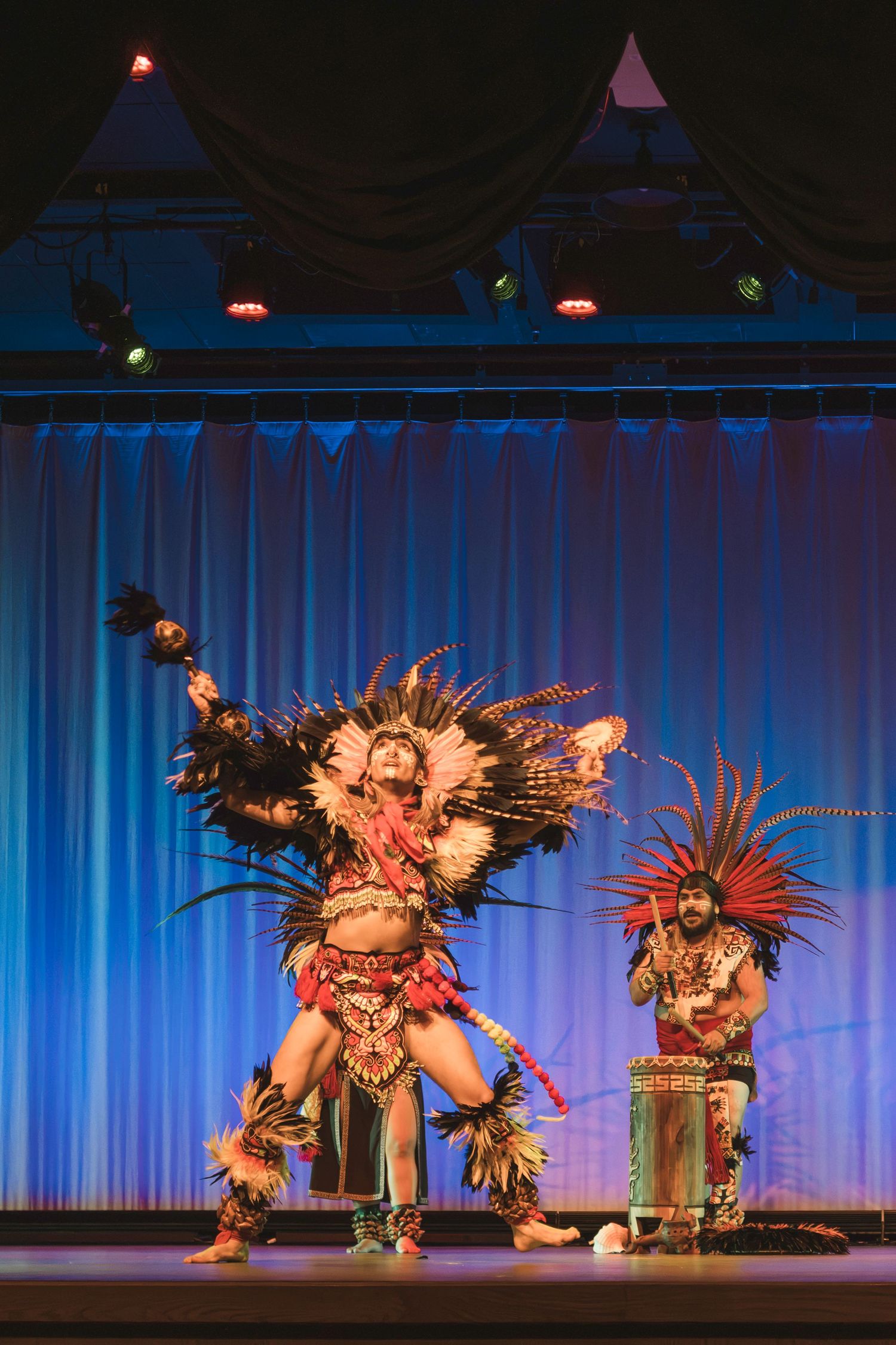 Two performers in elaborate Aztec ceremonial costumes with feathered headdresses dancing on a theater stage.