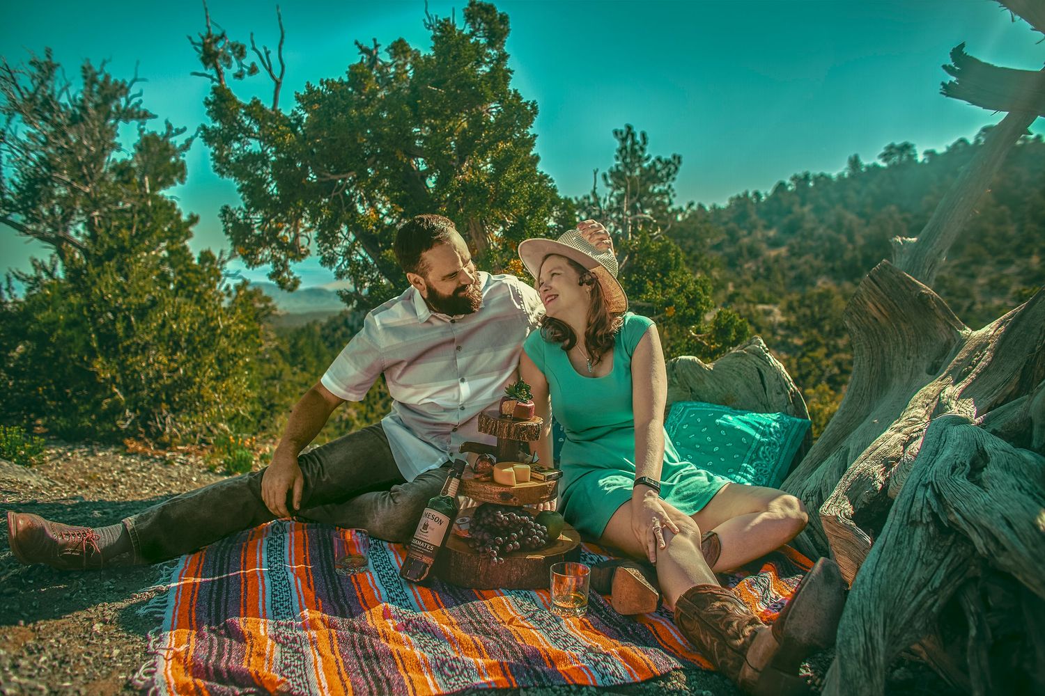Couple enjoying a romantic outdoor picnic on a colorful blanket surrounded by trees on a sunny day.