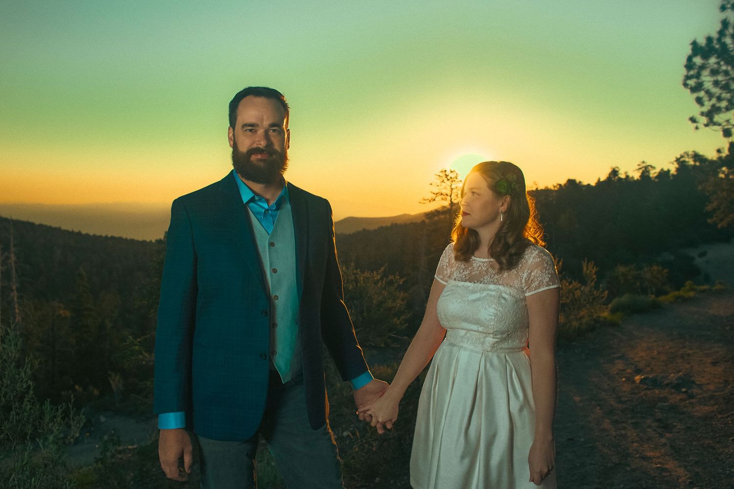 Couple in wedding attire holding hands at sunset with scenic mountain backdrop and vibrant sky.