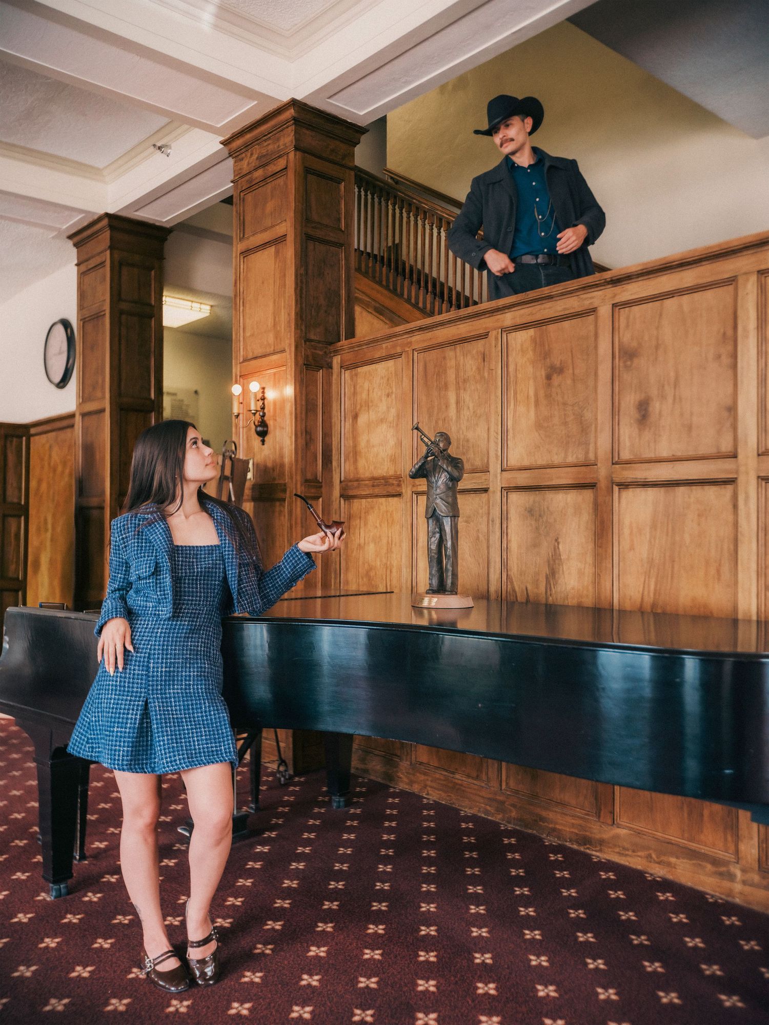 Woman in blue dress stands by piano as man in cowboy hat looks down from balcony in elegant wood-paneled lobby.
