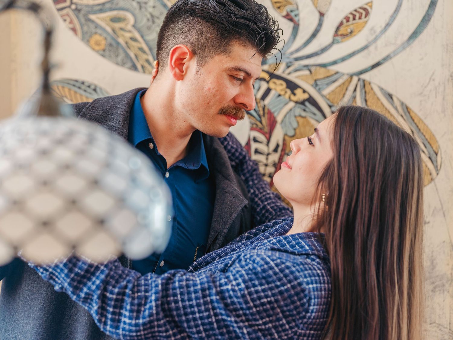 A romantic couple sharing an intimate moment, gazing into each other's eyes against a floral patterned wall.