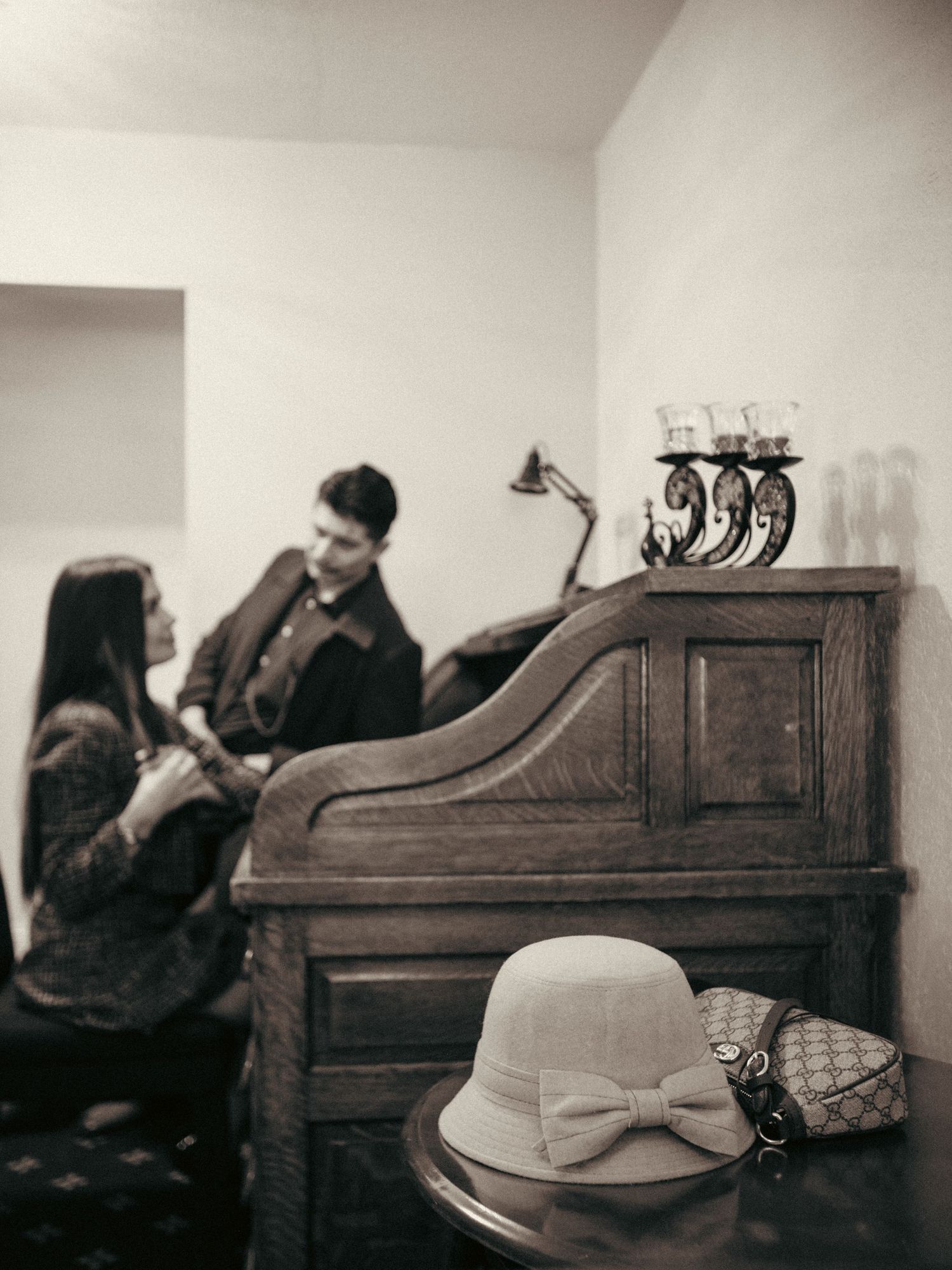 Vintage sepia photo of a couple near an antique upright piano, with a cloche hat and purse on a chair in the foreground.