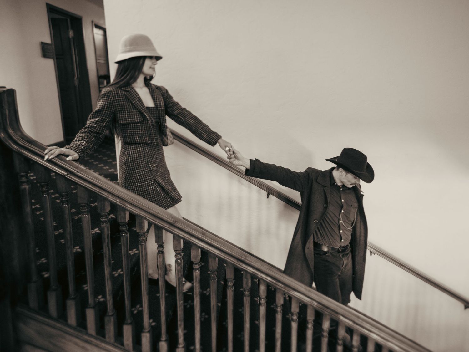 Two people in vintage western attire hold hands on a wooden staircase in a sepia-toned photograph.