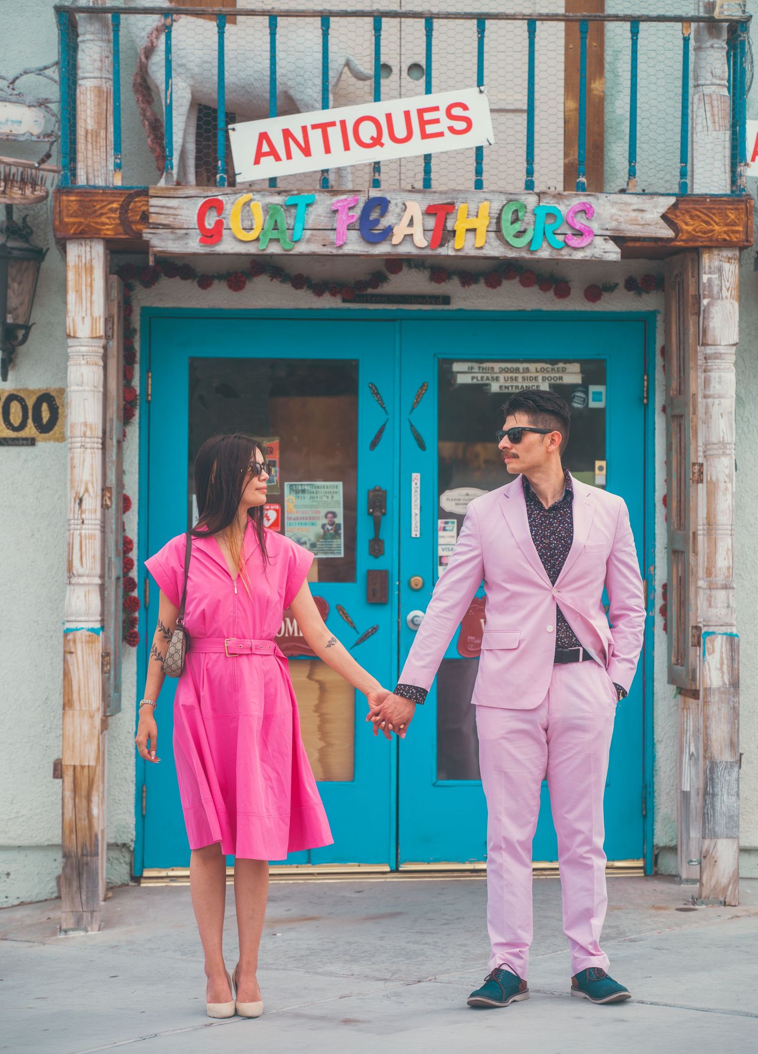 Couple in matching pink outfits holding hands outside a colorful antiques shop called Quit Feathers.