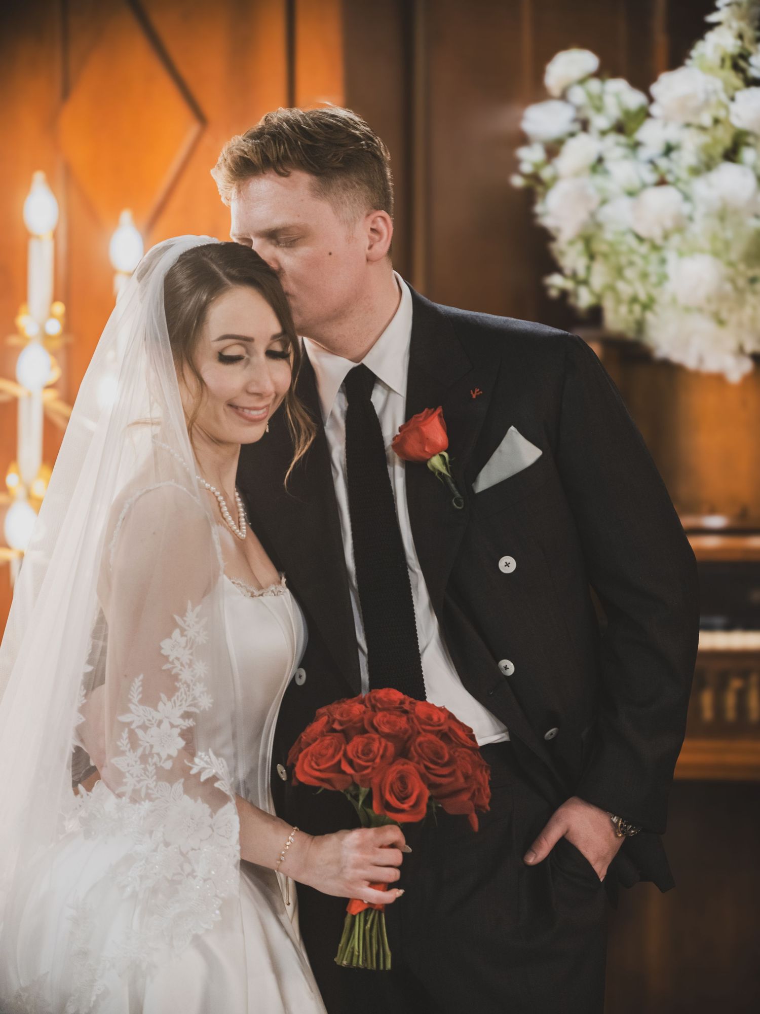 Bride and groom share a tender kiss during wedding ceremony, bride holds red rose bouquet wearing lace veil and white gown.
