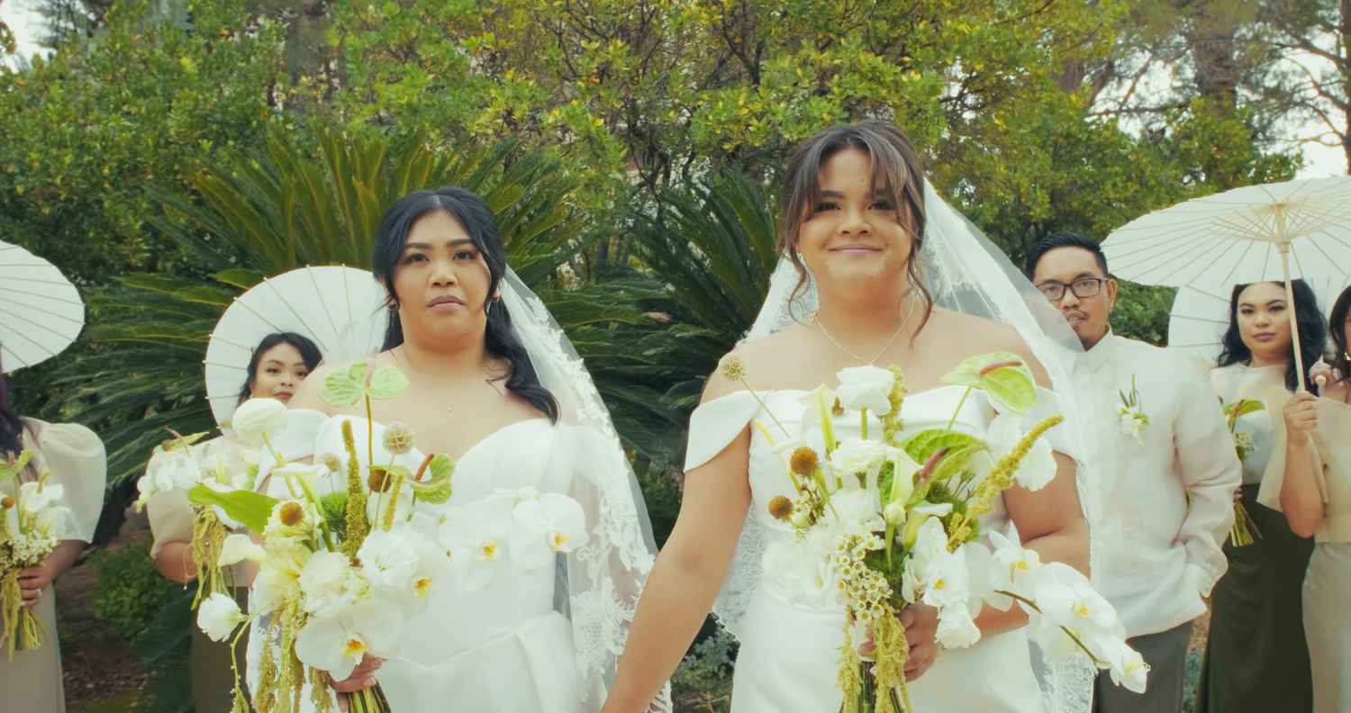 Two brides in white wedding gowns and veils walk together holding floral bouquets outdoors.
