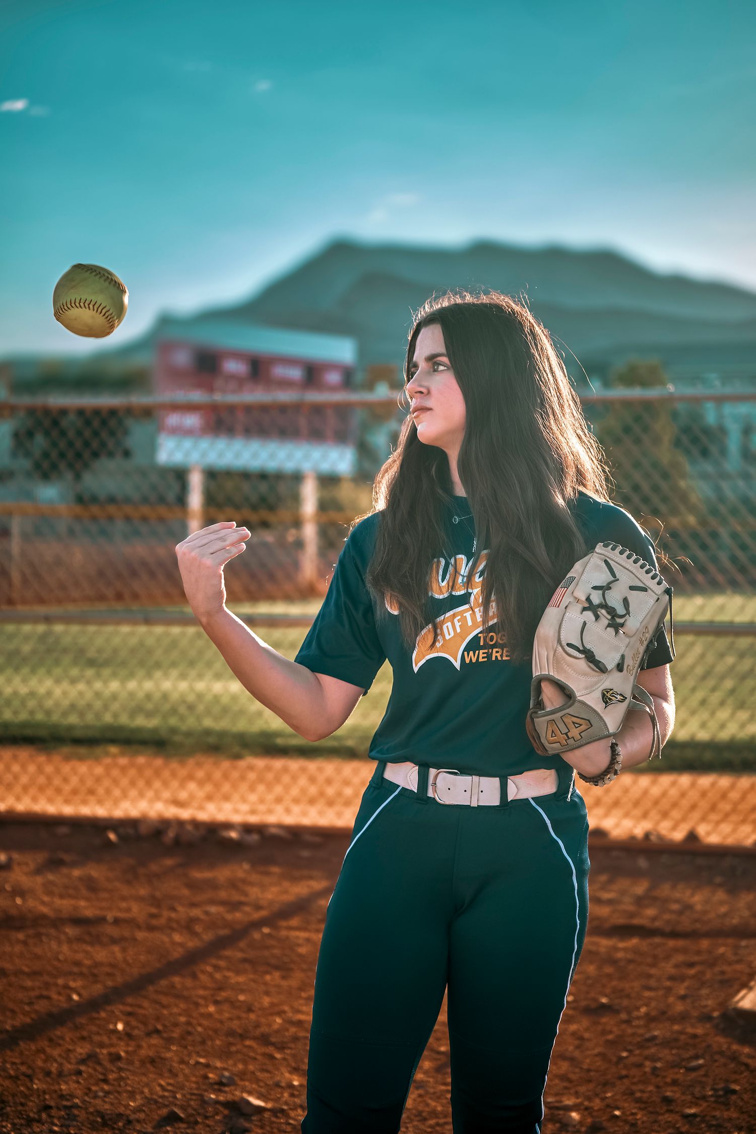 Young female softball player in green uniform tossing a ball in the air while holding a glove on a dirt field.