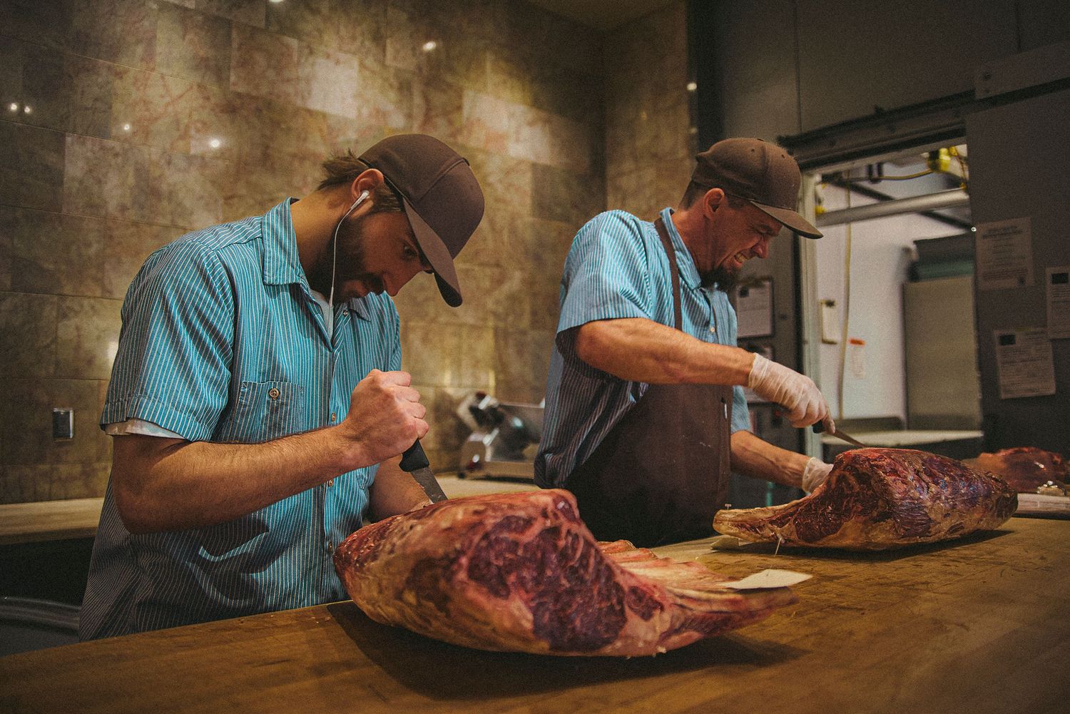 Two butchers in blue shirts and caps expertly carve large cuts of aged beef on a wooden counter.