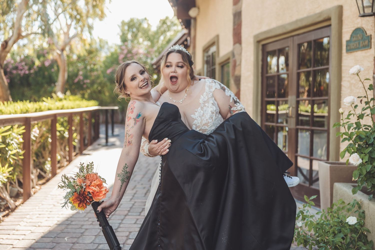 Two brides laughing as one carries the other on a wooden walkway at wedding.