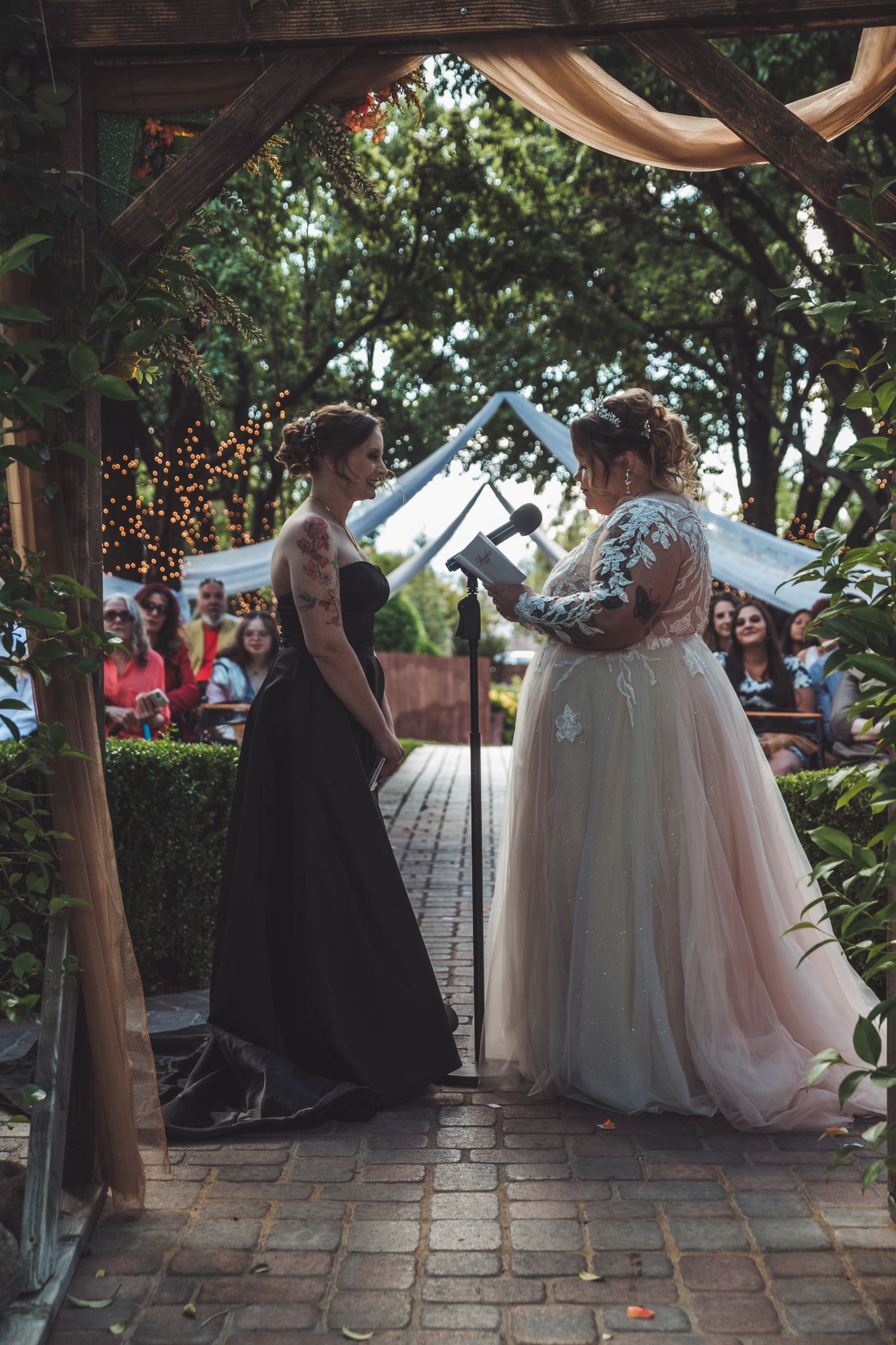 Two brides exchange vows under wooden arch at outdoor garden wedding ceremony.