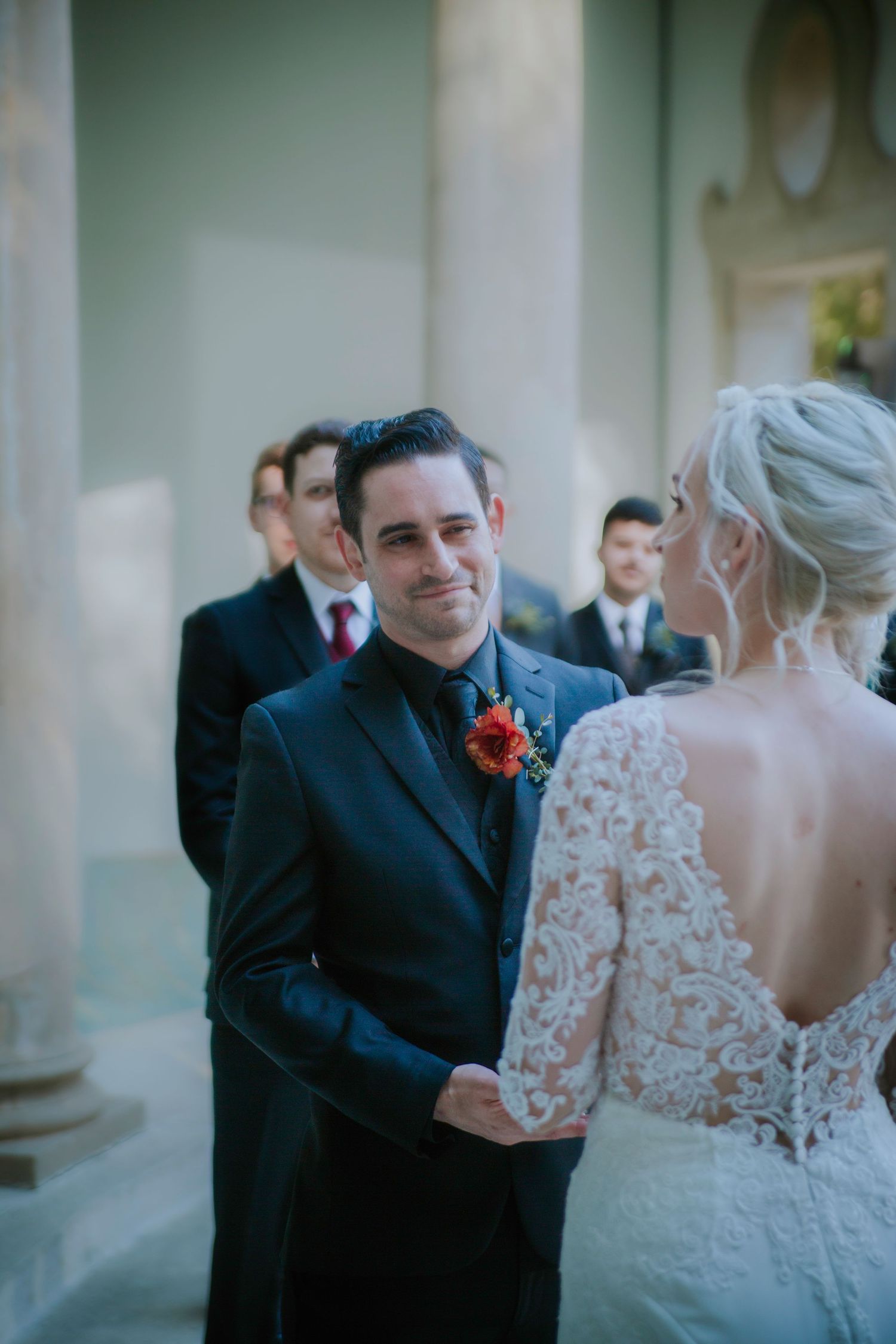 Emotional groom in black suit smiles at bride during wedding ceremony with groomsmen standing in background.