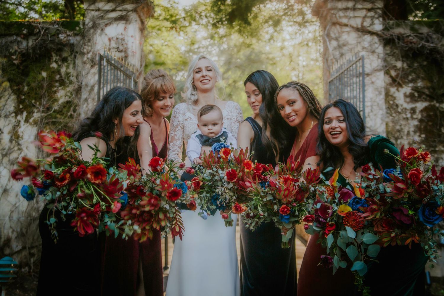 Bride in white lace gown poses with bridesmaids in jewel-toned dresses holding vibrant red and blue floral bouquets.