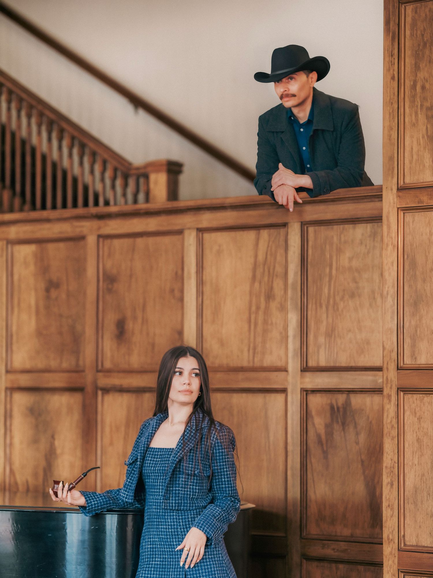 Man in black cowboy hat leaning over wooden balcony railing above woman in blue plaid blazer holding glasses.