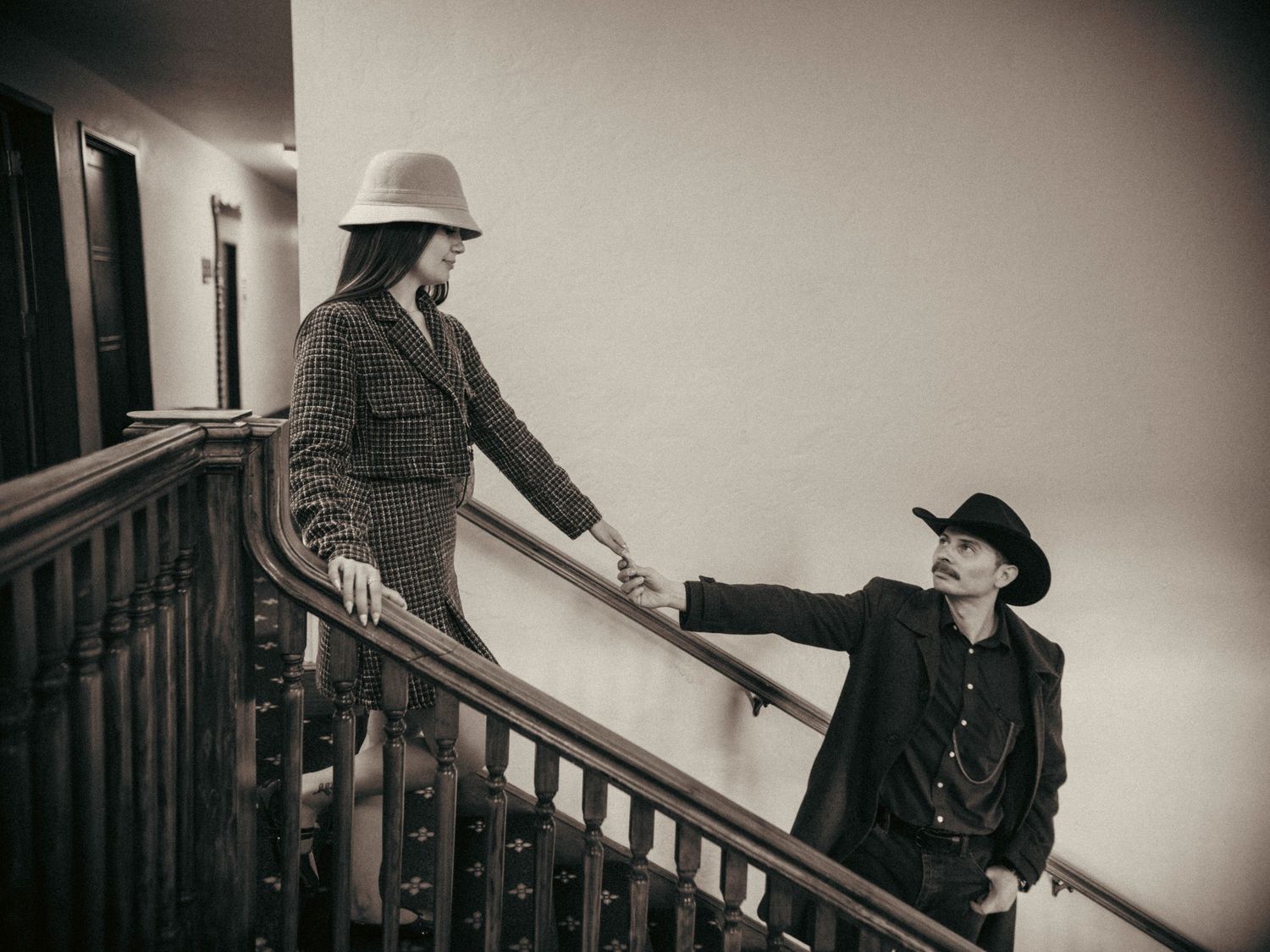 A couple in western hats hold hands on a staircase in a sepia-toned romantic photo.