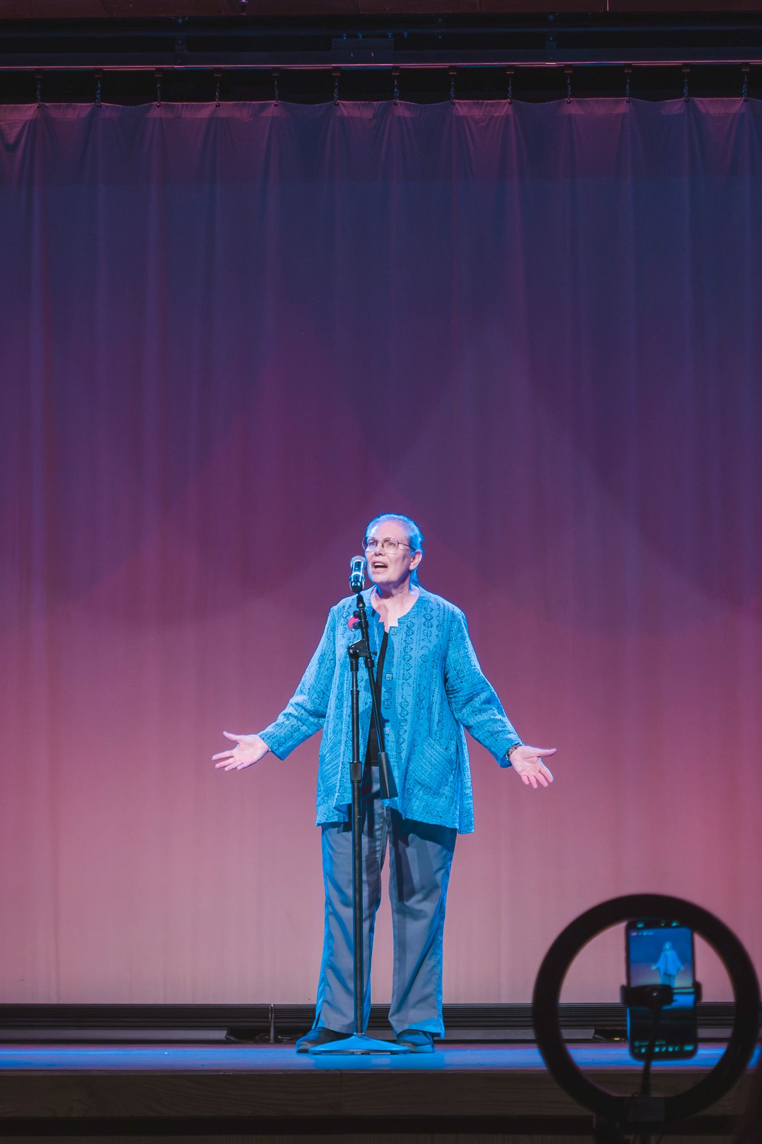 Performer in blue outfit stands at microphone on stage with arms outstretched, pink and purple curtain backdrop.