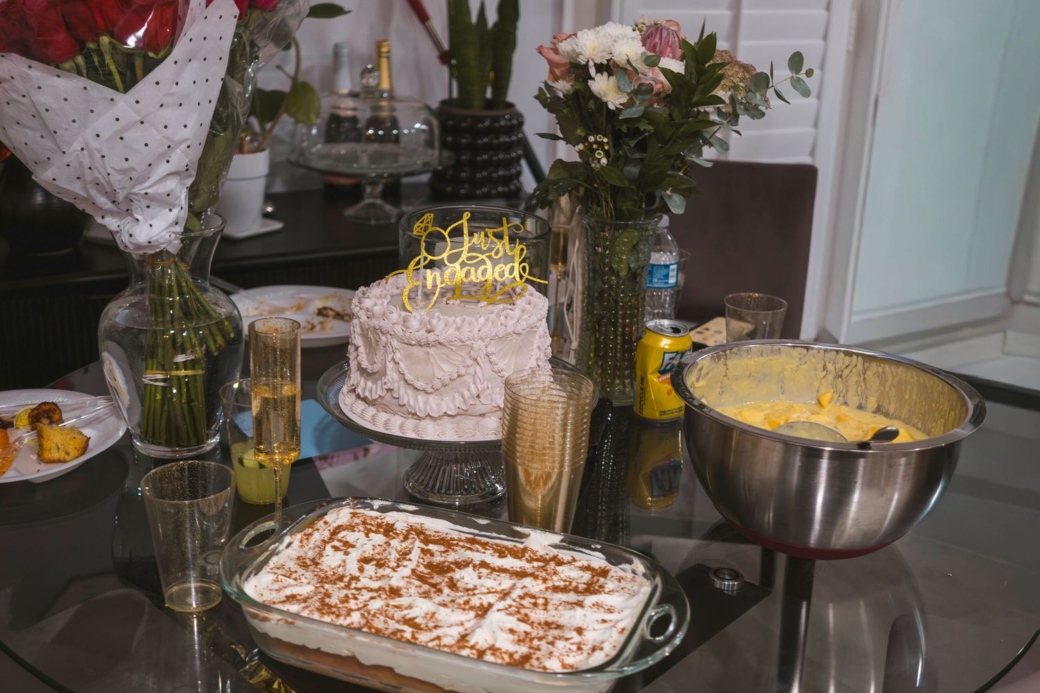 Birthday celebration table with white layered cake, tiramisu dessert, mixing bowl, floral arrangements, and drinks on glass table.
