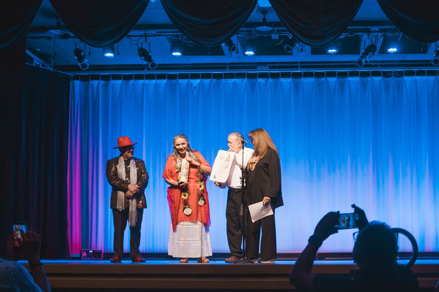 Four people on a blue-curtained stage during an award ceremony, one holding a certificate.