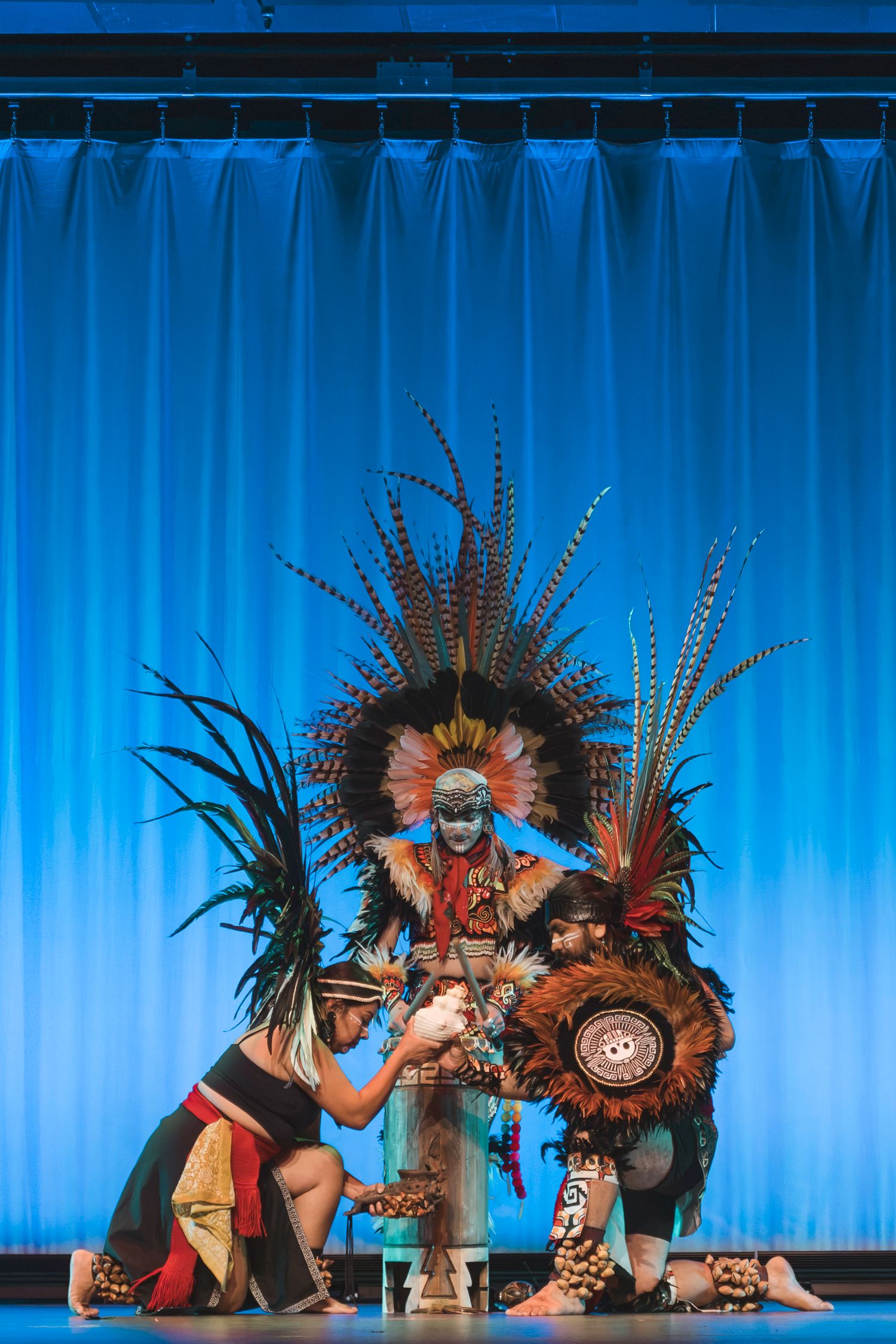 Three performers in elaborate Aztec ceremonial costumes with feathered headdresses perform a ritual dance on a blue-lit stage.