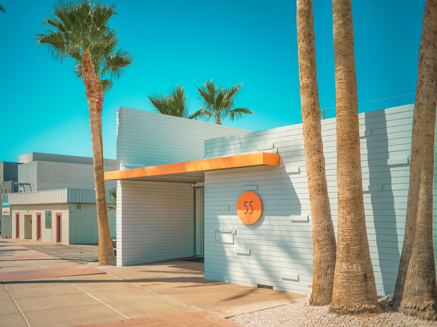 Retro mid-century modern building with white walls, orange accents, and tall palm trees under a bright blue sky.