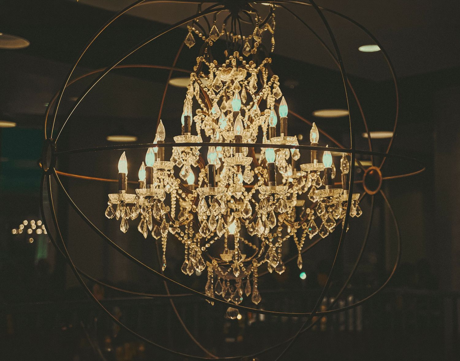 Ornate crystal chandelier with candle lights encased in a large metal orb sphere, hanging in a dimly lit elegant venue.
