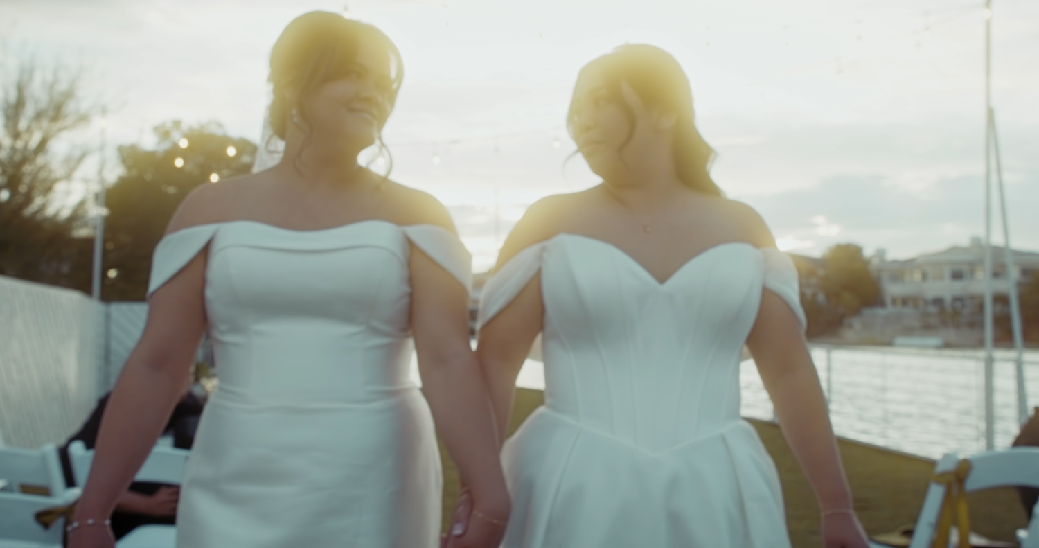 Two brides in white off-shoulder gowns hold hands walking together near a waterfront at sunset.