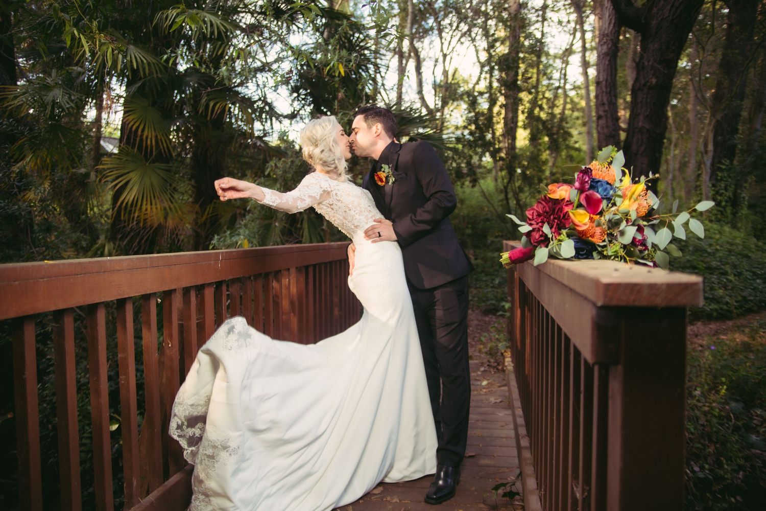 Bride in lace gown twirls while kissing groom on wooden bridge with colorful bridal bouquet resting on railing.