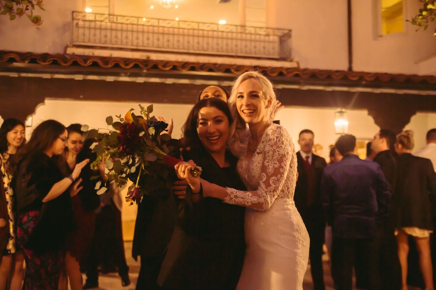 Smiling bride in lace gown hugging a guest while holding a bouquet at outdoor wedding reception.