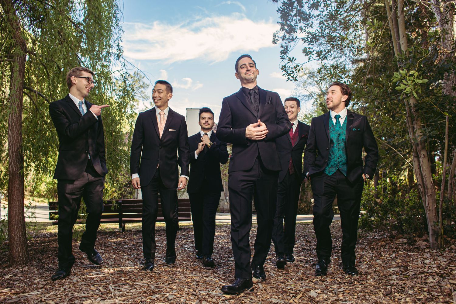 Groomsmen in dark suits posing outdoors with trees and blue sky background.
