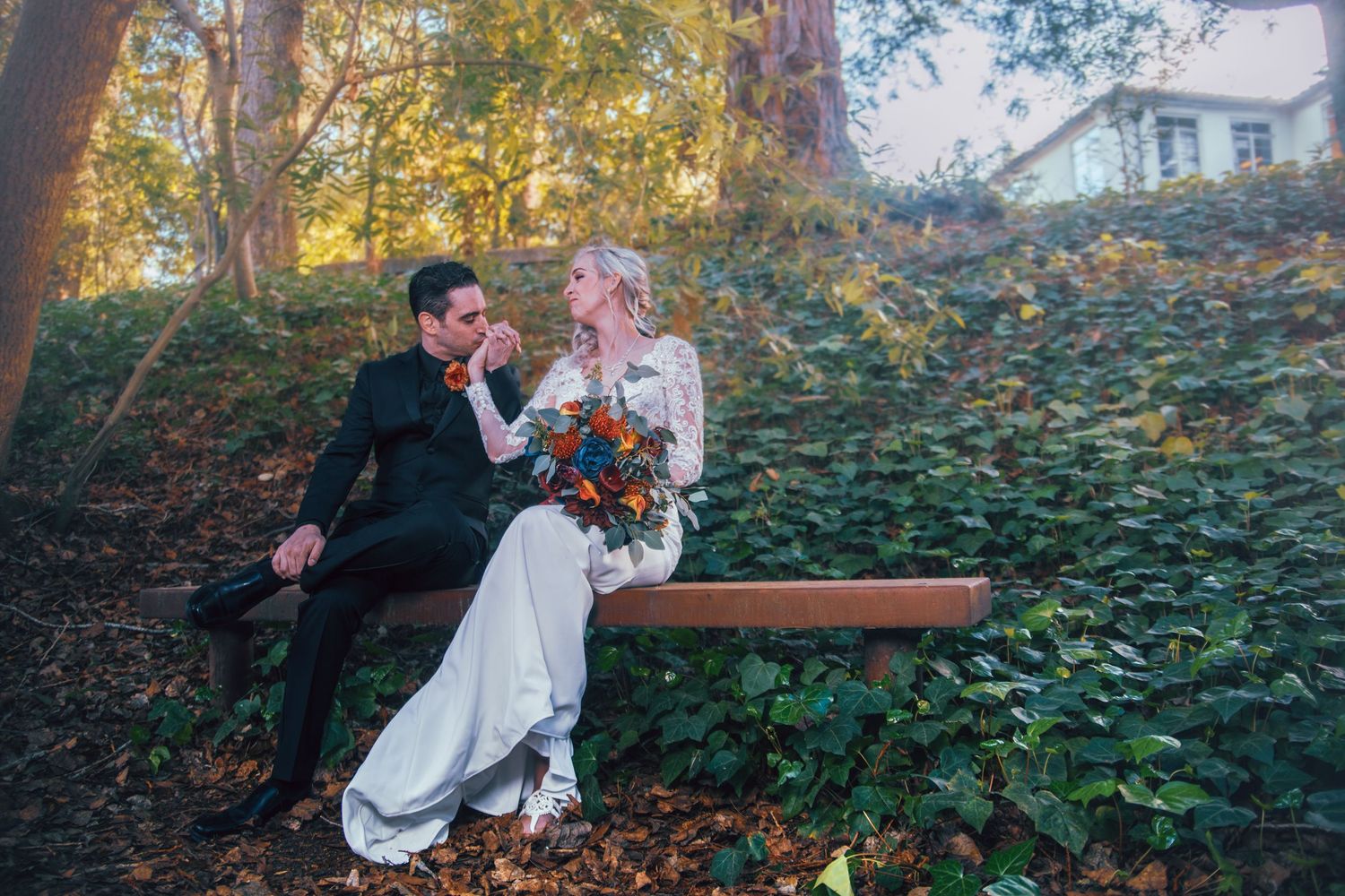A bride and groom sit on a wooden bench surrounded by ivy and autumn trees, sharing a romantic moment on their wedding day.