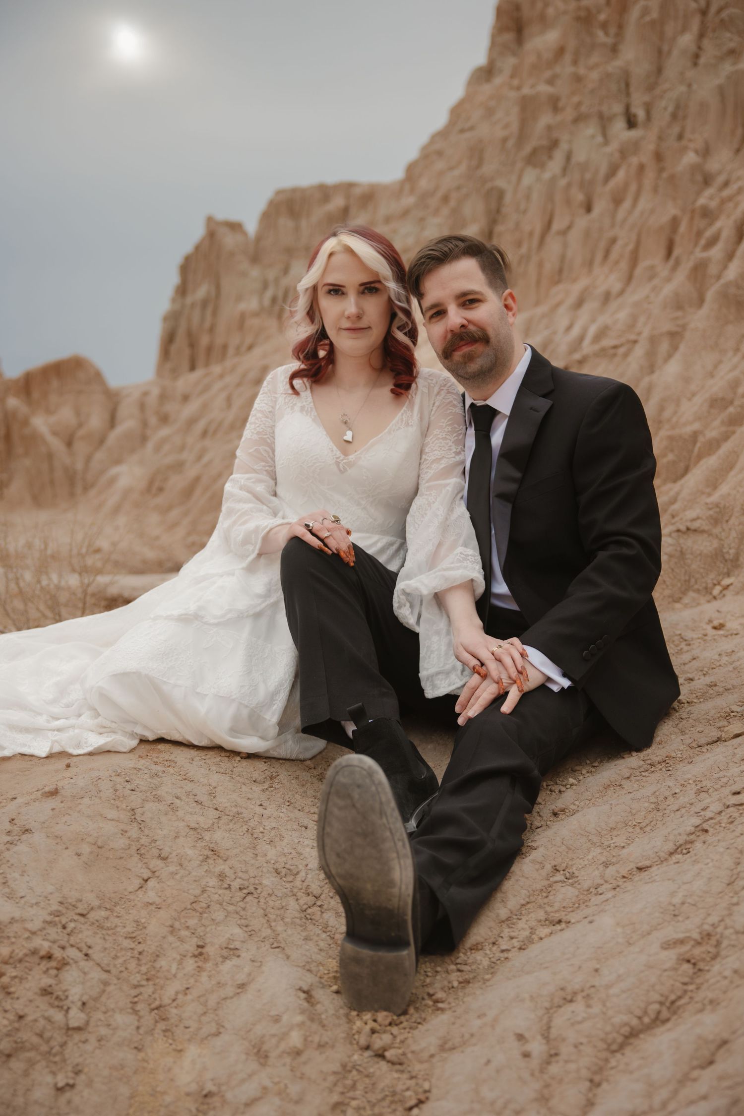 Bride and groom sit together on sandy desert terrain holding hands in romantic wedding portrait.