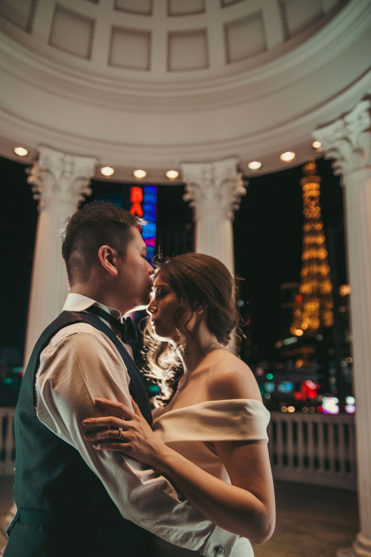 Couple sharing romantic moment under domed rotunda with Las Vegas Strip lights glowing in background at night.