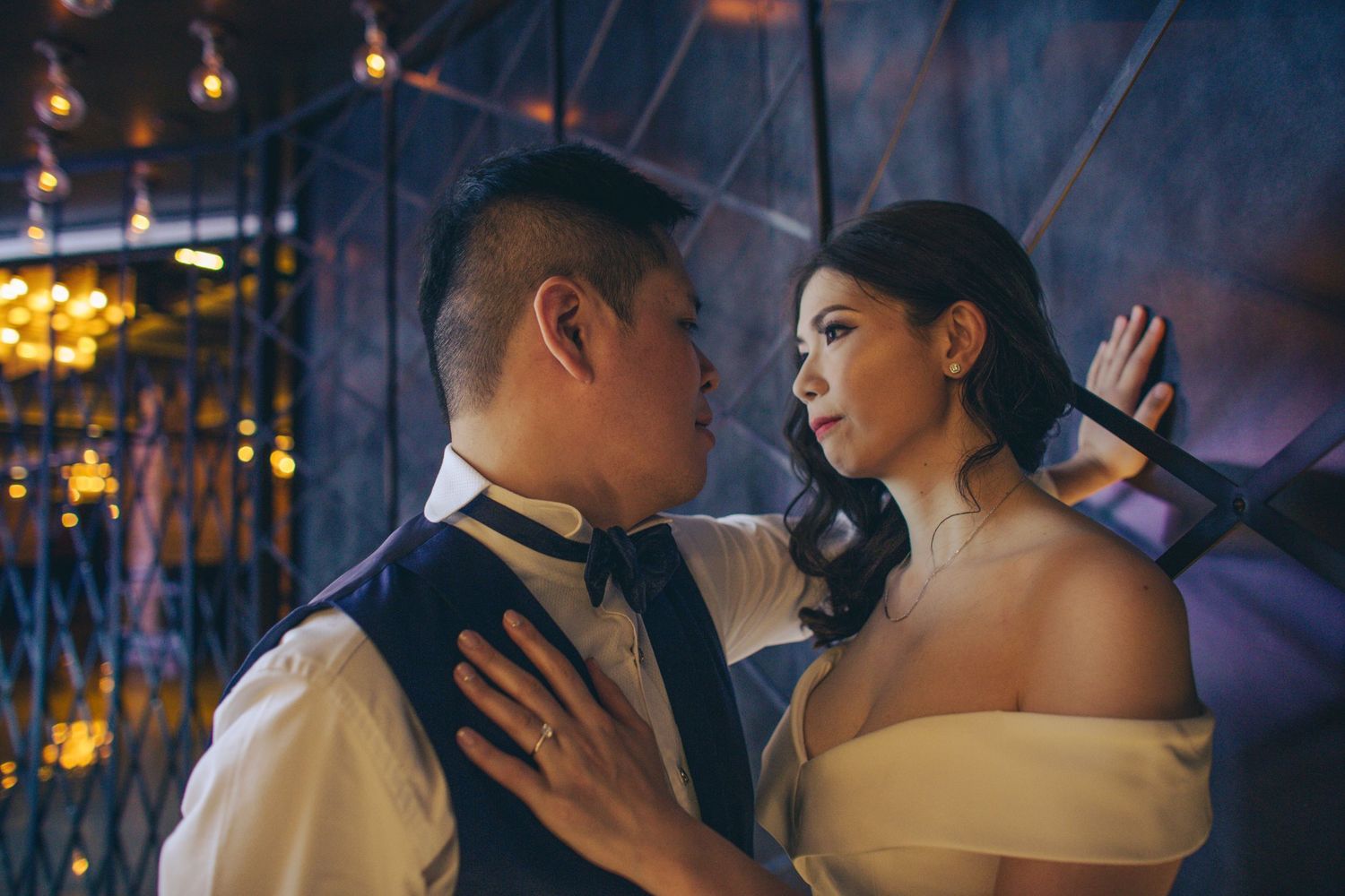 Bride and groom sharing an intimate gaze indoors with warm bokeh lights in the background during wedding shoot.